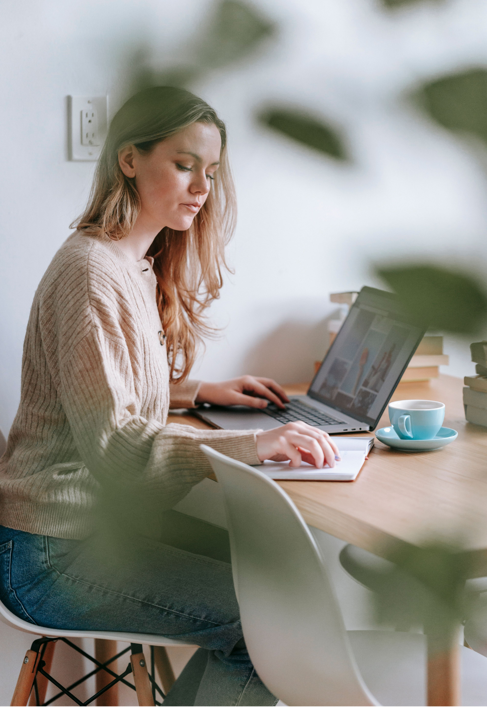 A woman with long, wavy hair in a beige sweater sitting at a wooden table working on a laptop, with a notepad and a cup nearby. Green leaves partially obscure the view in the foreground.