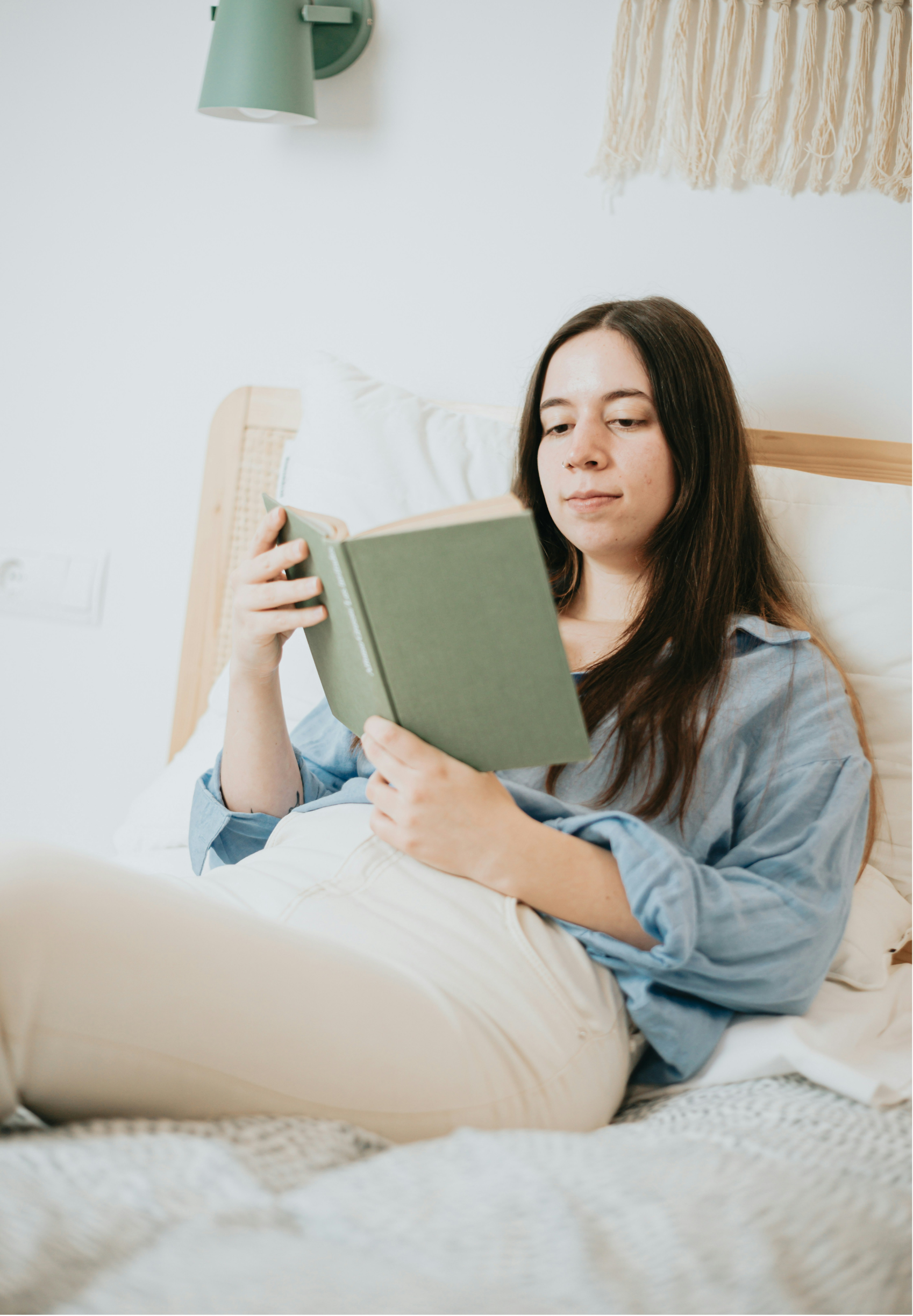 A young woman with long brown hair is lounging in bed, reading a green hardcover book. She is wearing a light blue denim shirt and beige pants, and the setting is a cozy bedroom with a white wall, a wooden bed frame, and decorative wall hangings.
