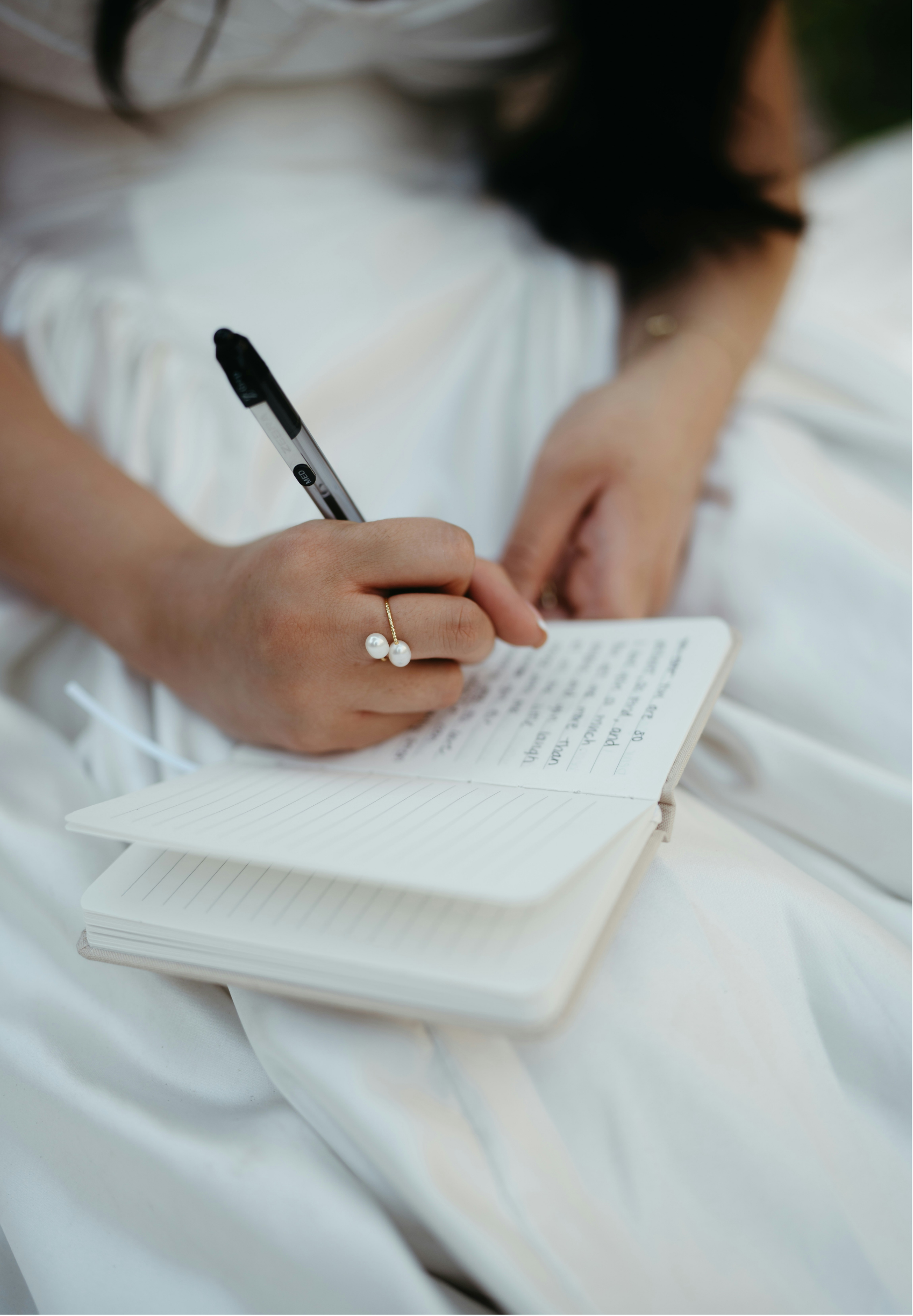 Person writing in a notebook with a pen, sitting on white fabric, wearing a ring with pearl earrings.