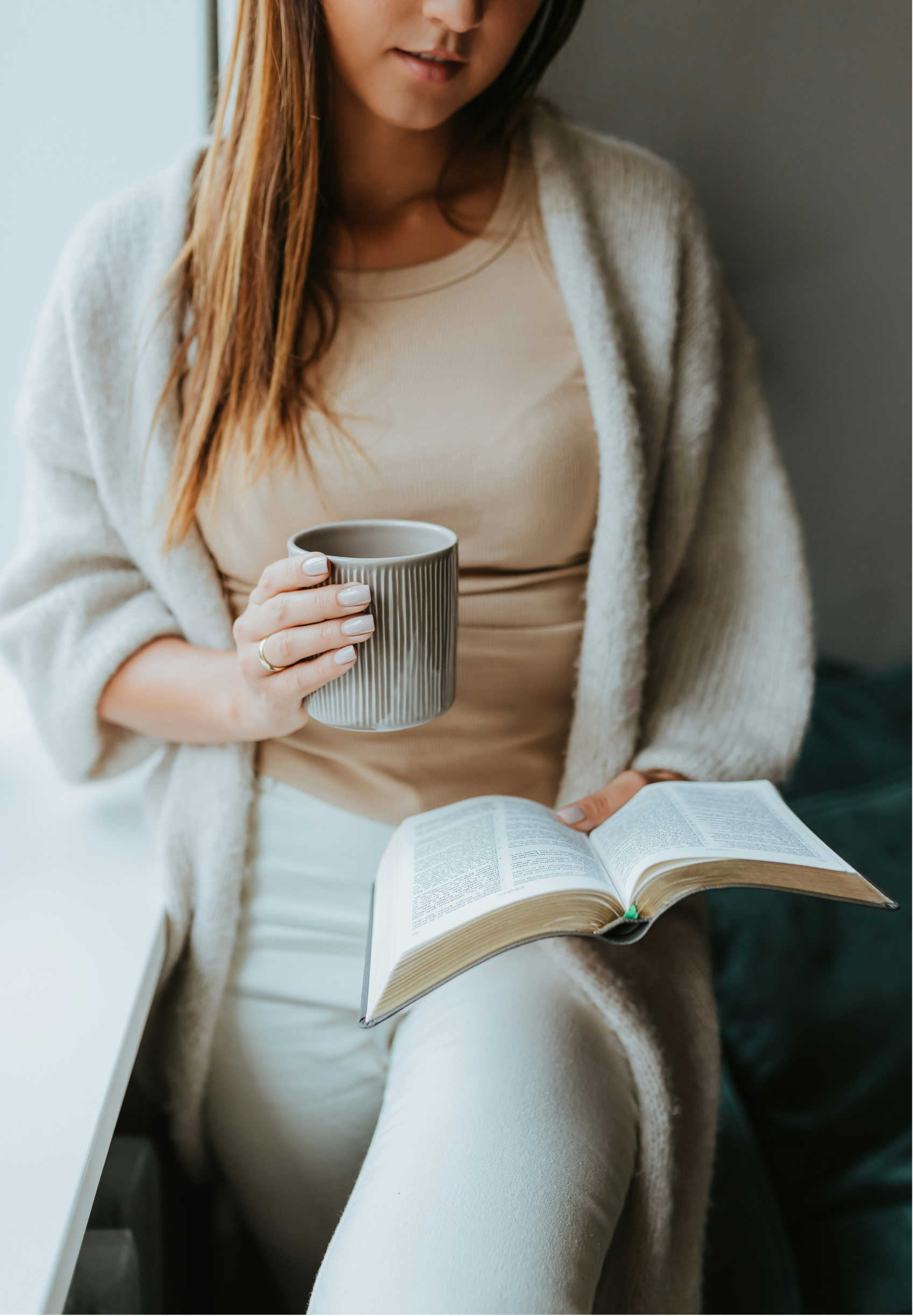 A woman with long reddish-brown hair, wearing a beige top and a cream-colored cardigan, holding a grayish striped mug in one hand and an open book in the other, sitting near a window.