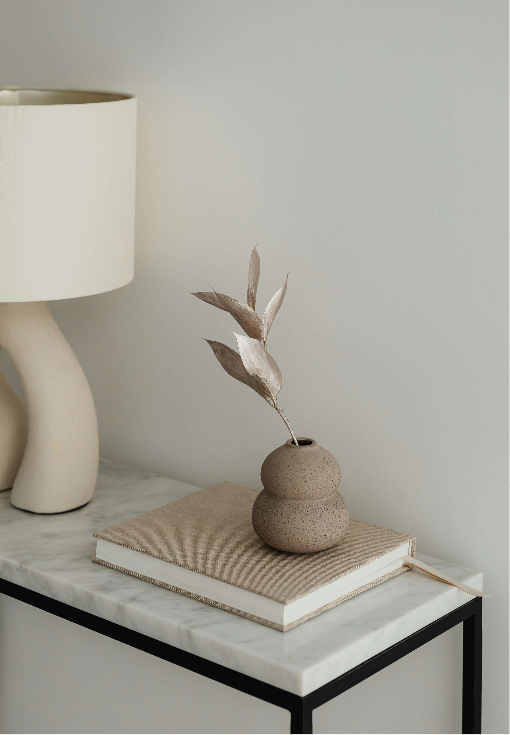 A minimalist interior scene with a small, round, beige clay vase holding dried, beige leaves, placed on a beige book on a white marble table with a black frame. To the left, a large off-white ceramic table lamp with a curved base and a cream shade is partially visible. The background is a plain, light-colored wall.