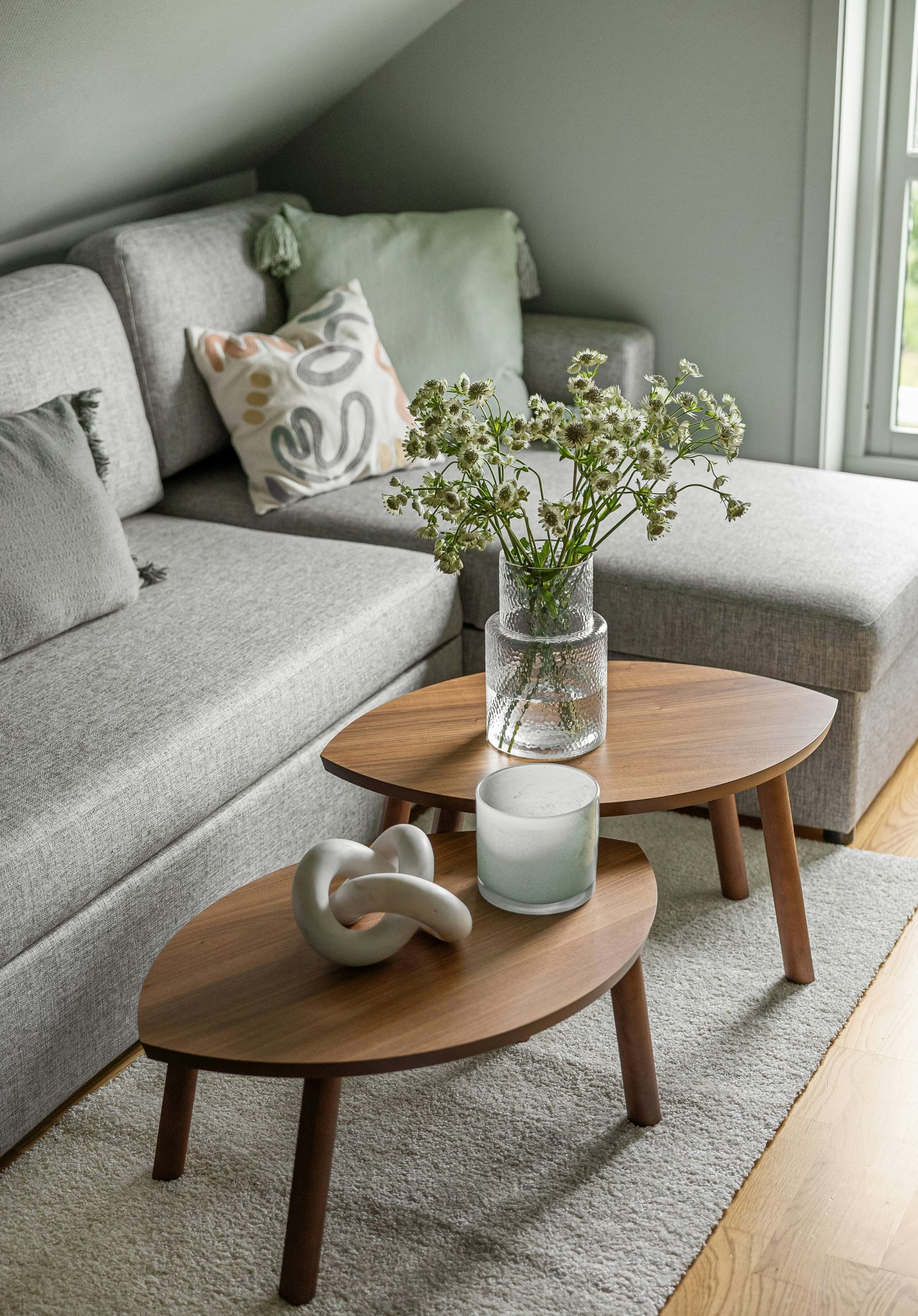 Living room with gray sectional sofa, decorative pillows, wooden coffee tables with vases, and a window with natural light.