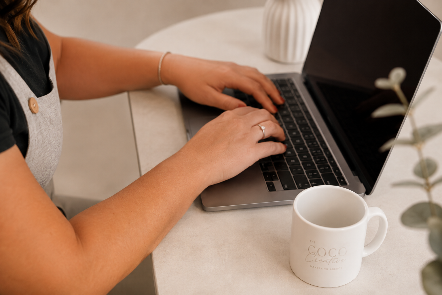 A woman typing on a laptop at a desk with a white coffee mug labeled 'The Goco Creative Marketing Agency' and a plant nearby.