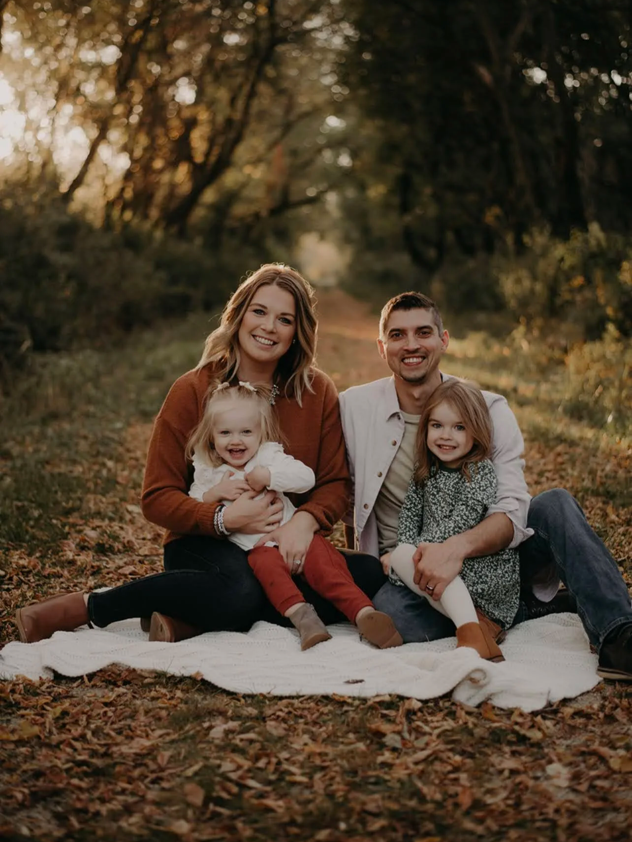 A family of four sitting on a blanket outdoors in an autumn forest, smiling at the camera.