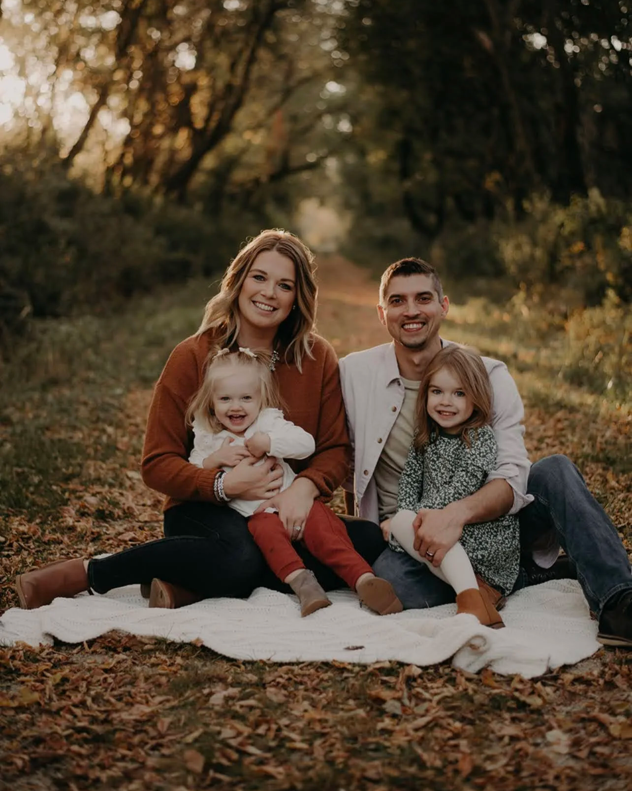 A family of four sitting on a blanket in a wooded area during sunset, smiling at the camera. The mother has shoulder-length hair and is holding a young girl with blonde hair. The father has short dark hair and is sitting next to a young girl with red hair. The ground is covered with fallen leaves.