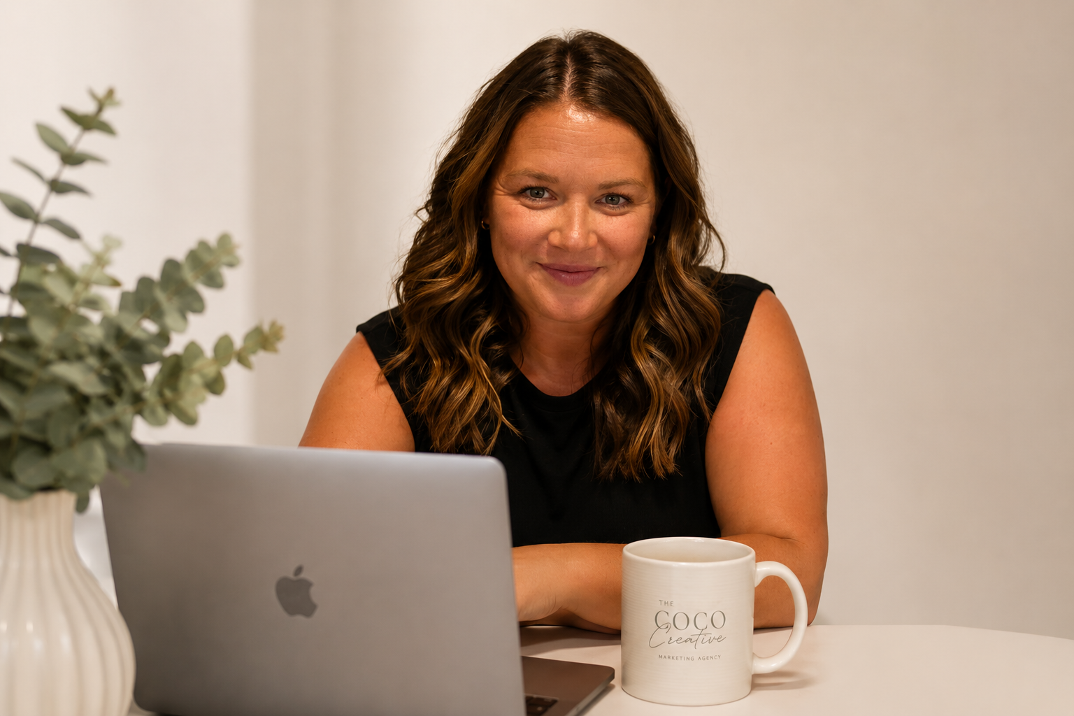 A woman with wavy brown hair smiling at the camera, sitting at a desk with a laptop, a white coffee mug that reads 'The GoCo Creative Marketing Agency,' and a potted plant in a beige vase.
