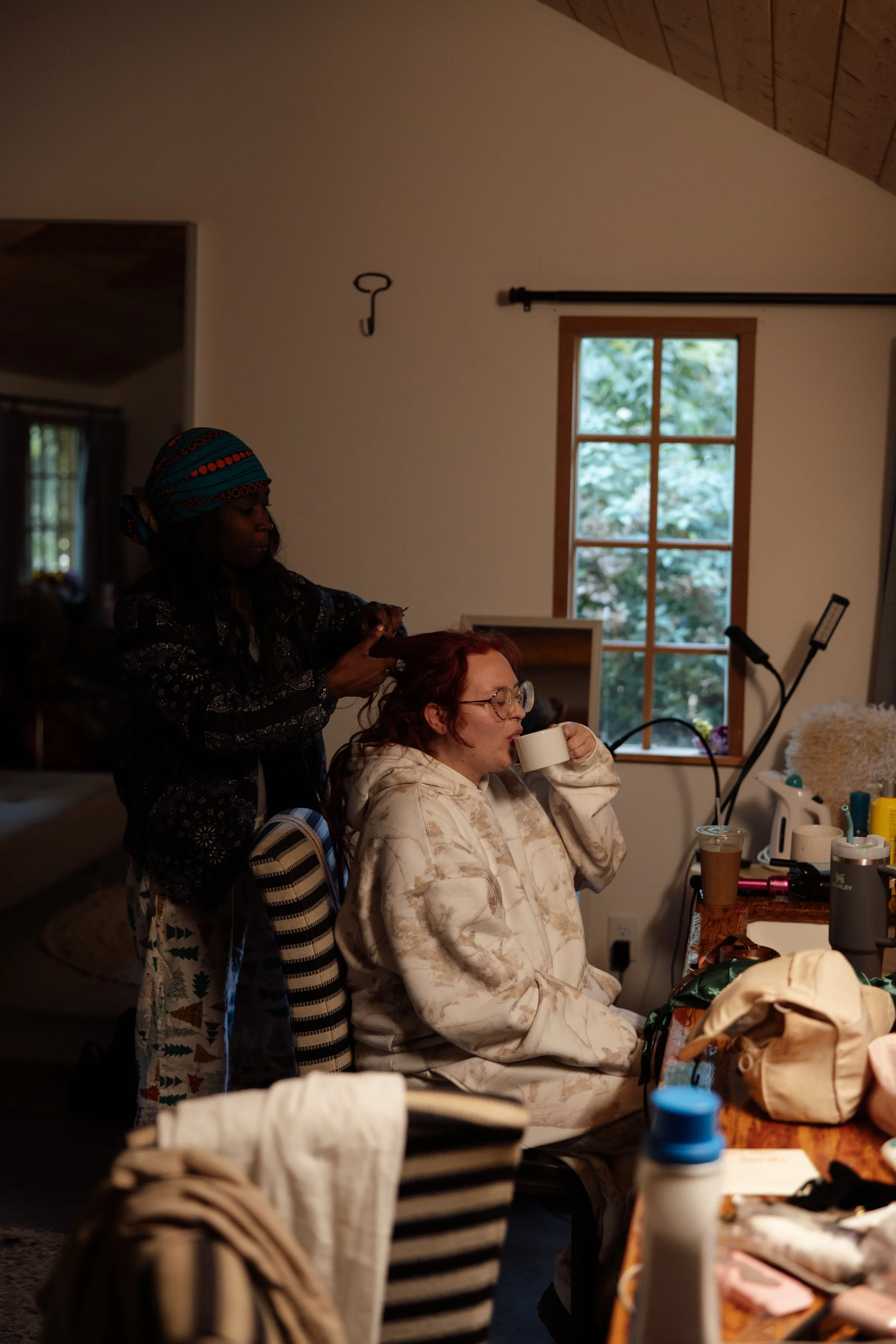 A woman with red hair and glasses sitting at a table, drinking from a mug, while another woman with dark skin and a colorful headscarf styles her hair in a well-lit room with a window showing trees outside.