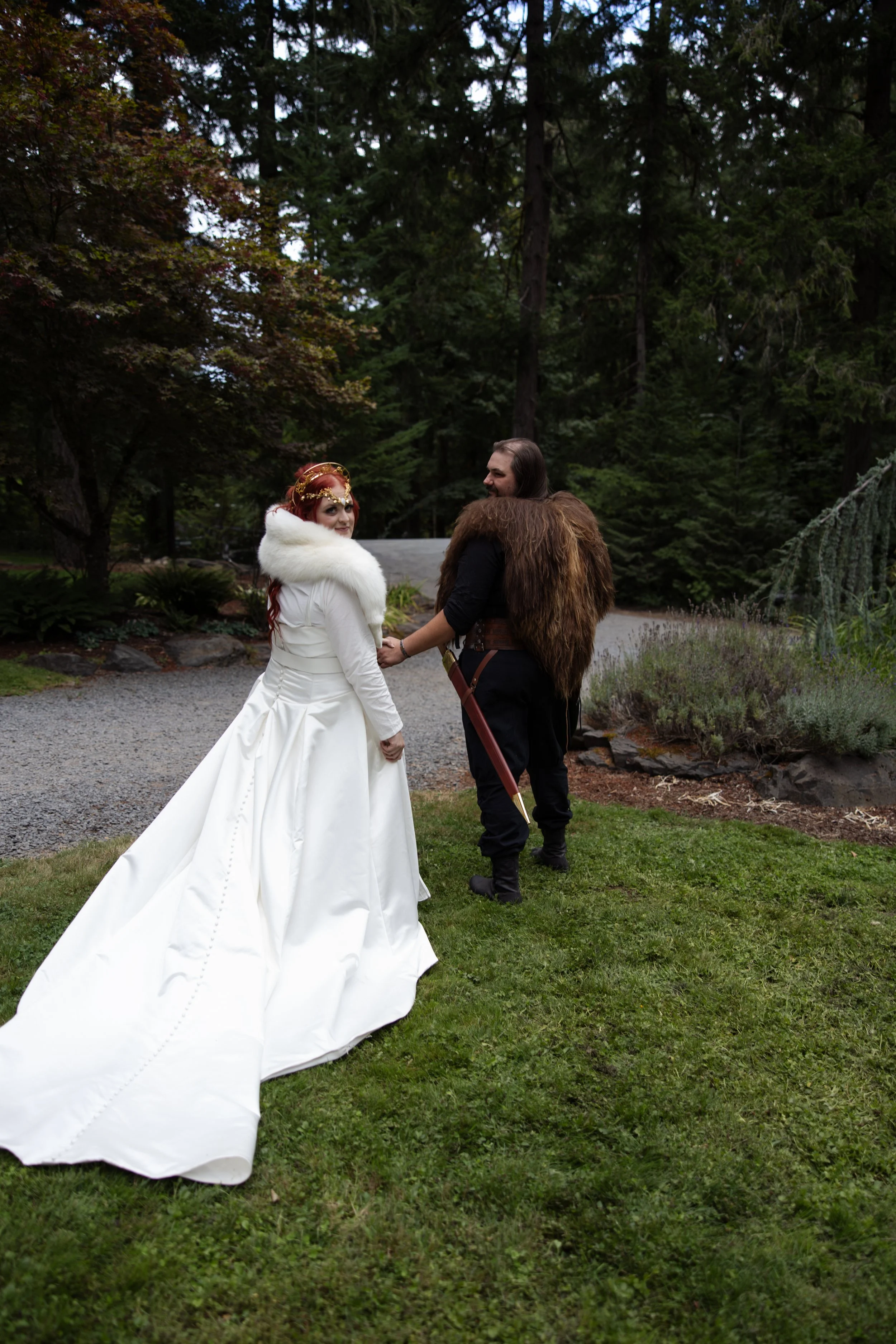 A bride and groom holding hands outdoors in a forested area during daytime. The bride is wearing a white gown with a long train and a fur stole, while the groom is dressed in medieval-style clothing with a fur cape and holding a sword.