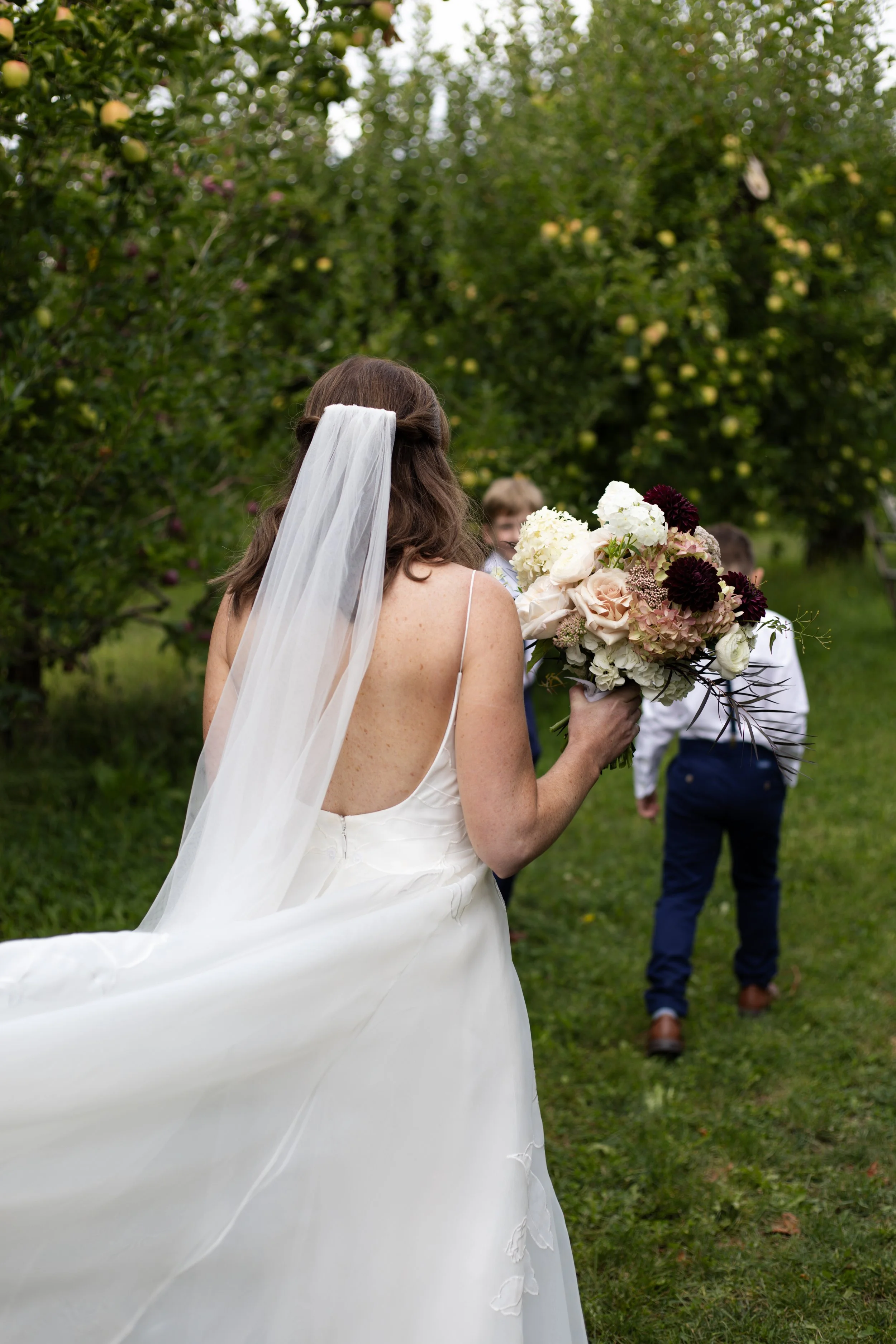 A woman wearing a white wedding dress and veil holds a large bouquet of flowers while walking outdoors in a garden, with a man in dress clothes and children in the background.