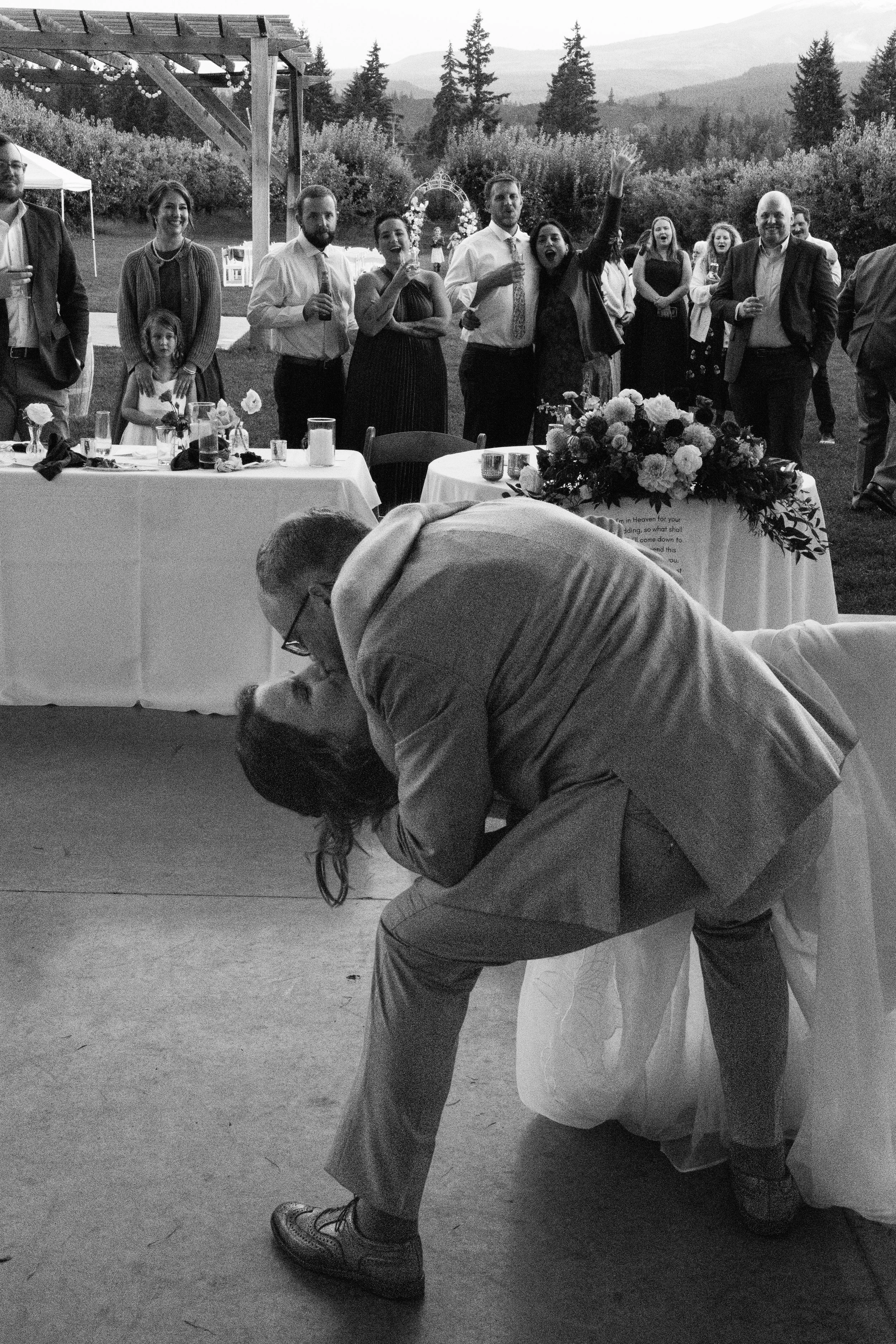A group of people at an outdoor celebration or wedding, with a man and woman dancing closely on the ground while others stand and watch, some holding drinks. There is a table with flowers and candles in the foreground, and trees and mountains in the 