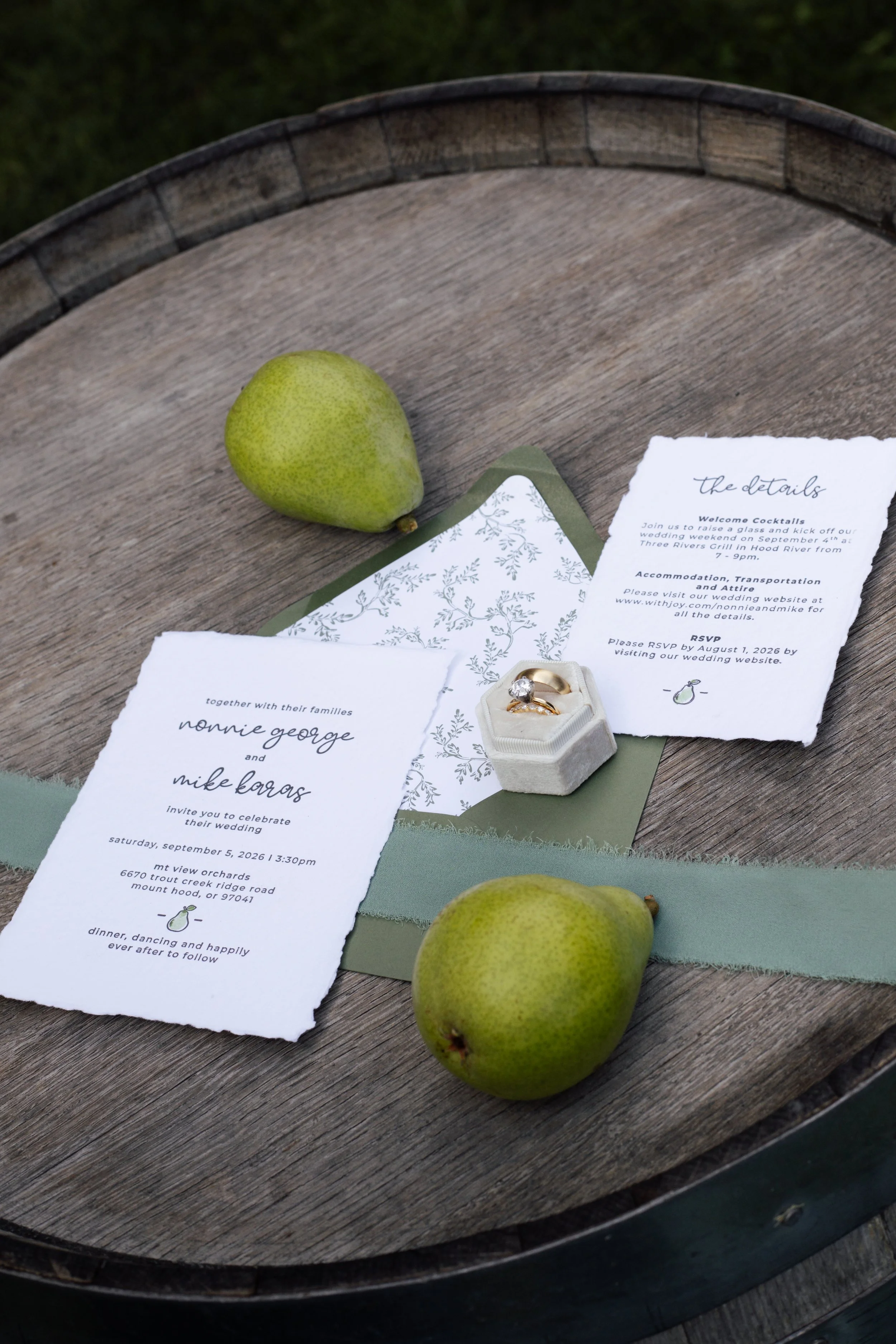 Green pears, wedding invitations, and a ring in a jewelry box on a wooden surface.