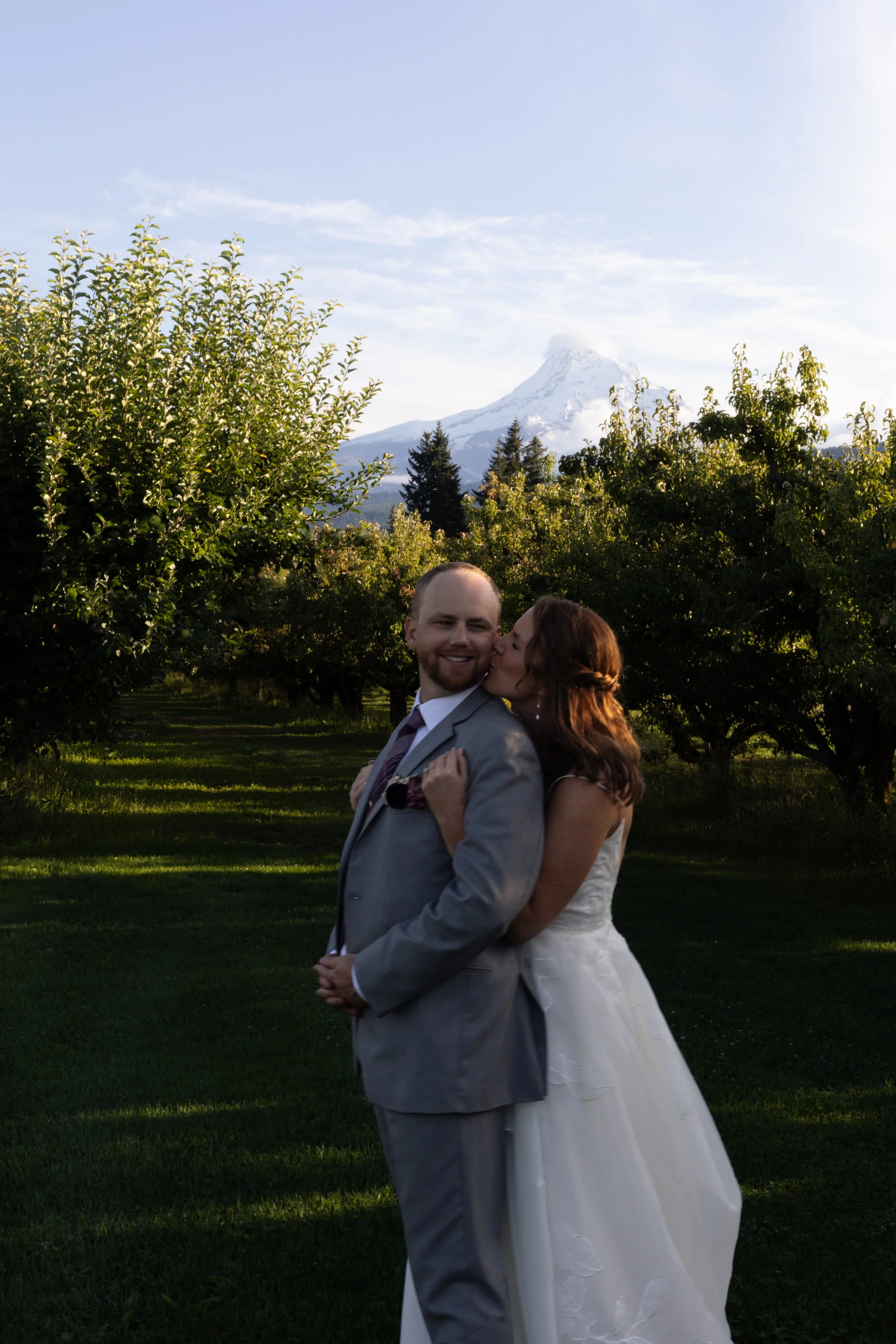 A newlywed couple in wedding attire, standing outdoors on a grassy area with trees, mountain, and snow-capped peak in the background. The woman is kissing the man on the cheek, and they are both smiling. The man is wearing a light gray suit and a tie