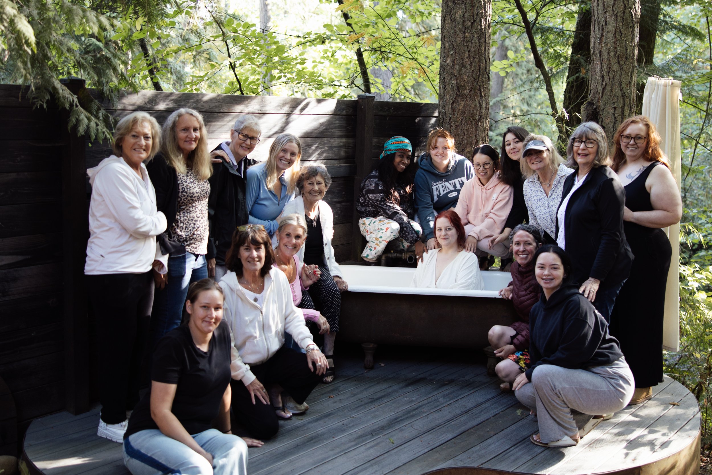 A diverse group of women and girls gathered outdoors next to a bathtub, surrounded by trees and greenery, smiling at the camera.