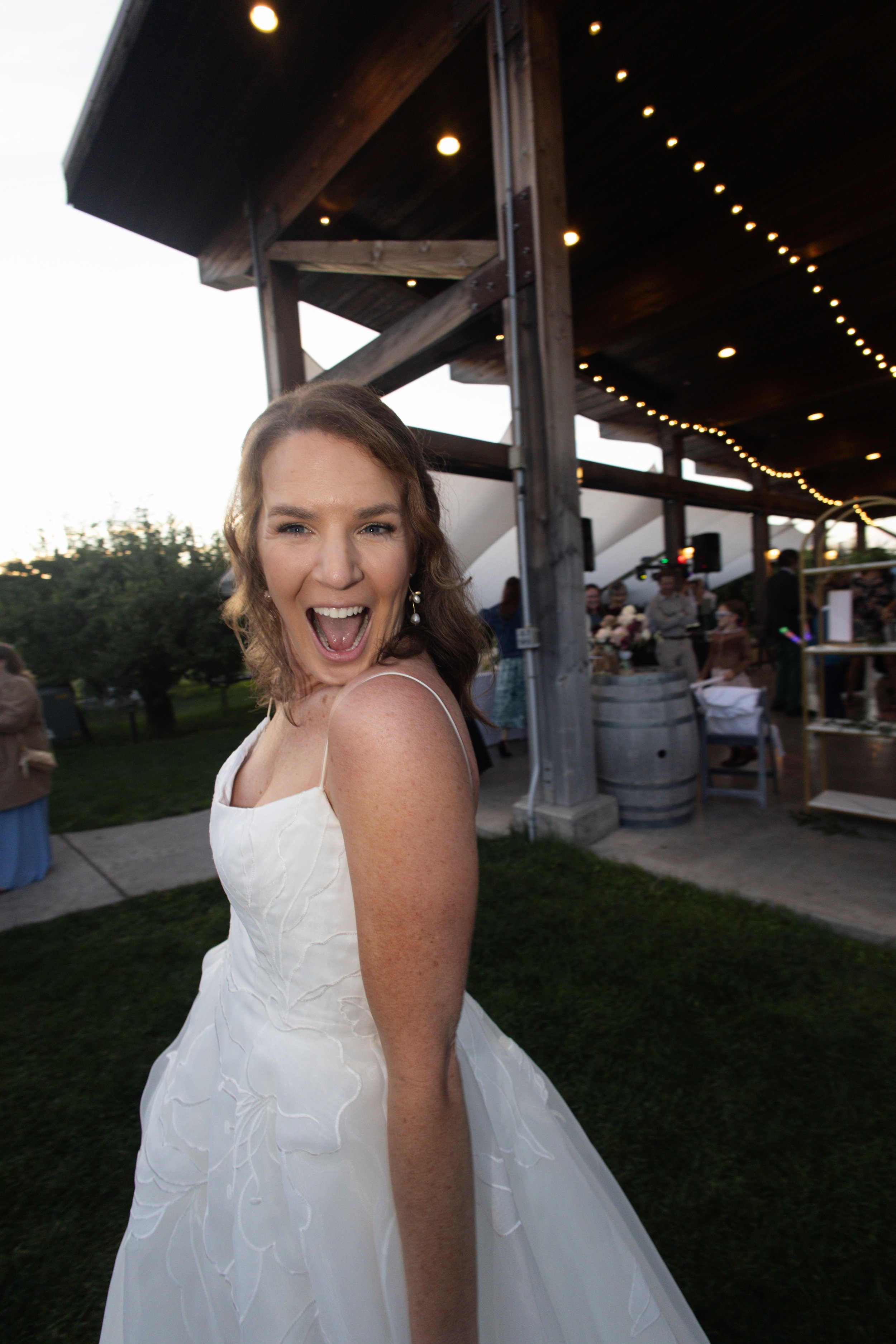 Woman in a white wedding dress joyfully smiling at an outdoor event during sunset, with outdoor string lights and guests in the background.