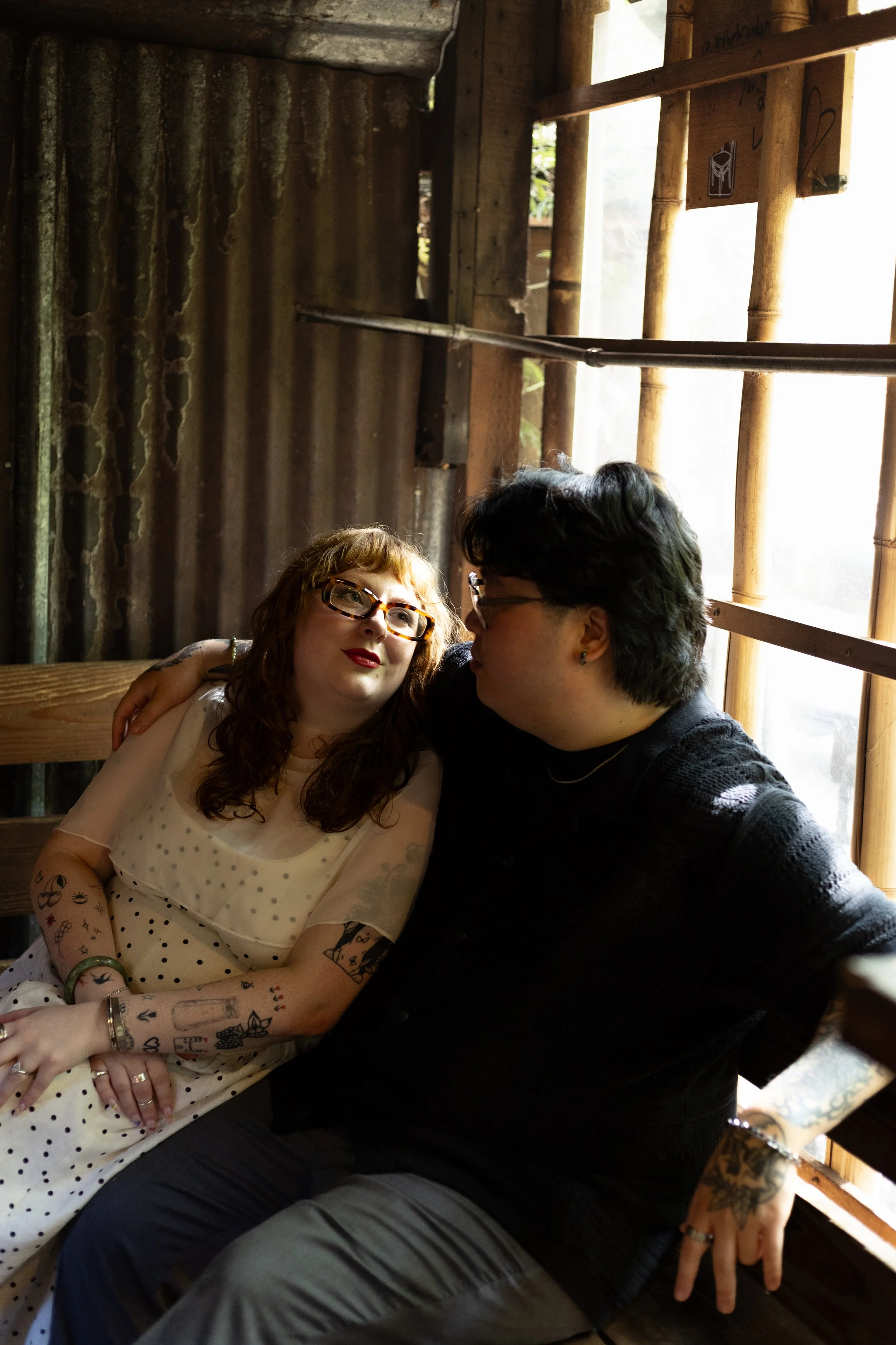 A woman with red hair, glasses, and tattoos on her arms, wearing a white polka dot dress, leaning on a man with dark hair, glasses, and tattoos on his arm, sitting together in a rustic, wooden setting with bamboo details, near a window with sunlight.