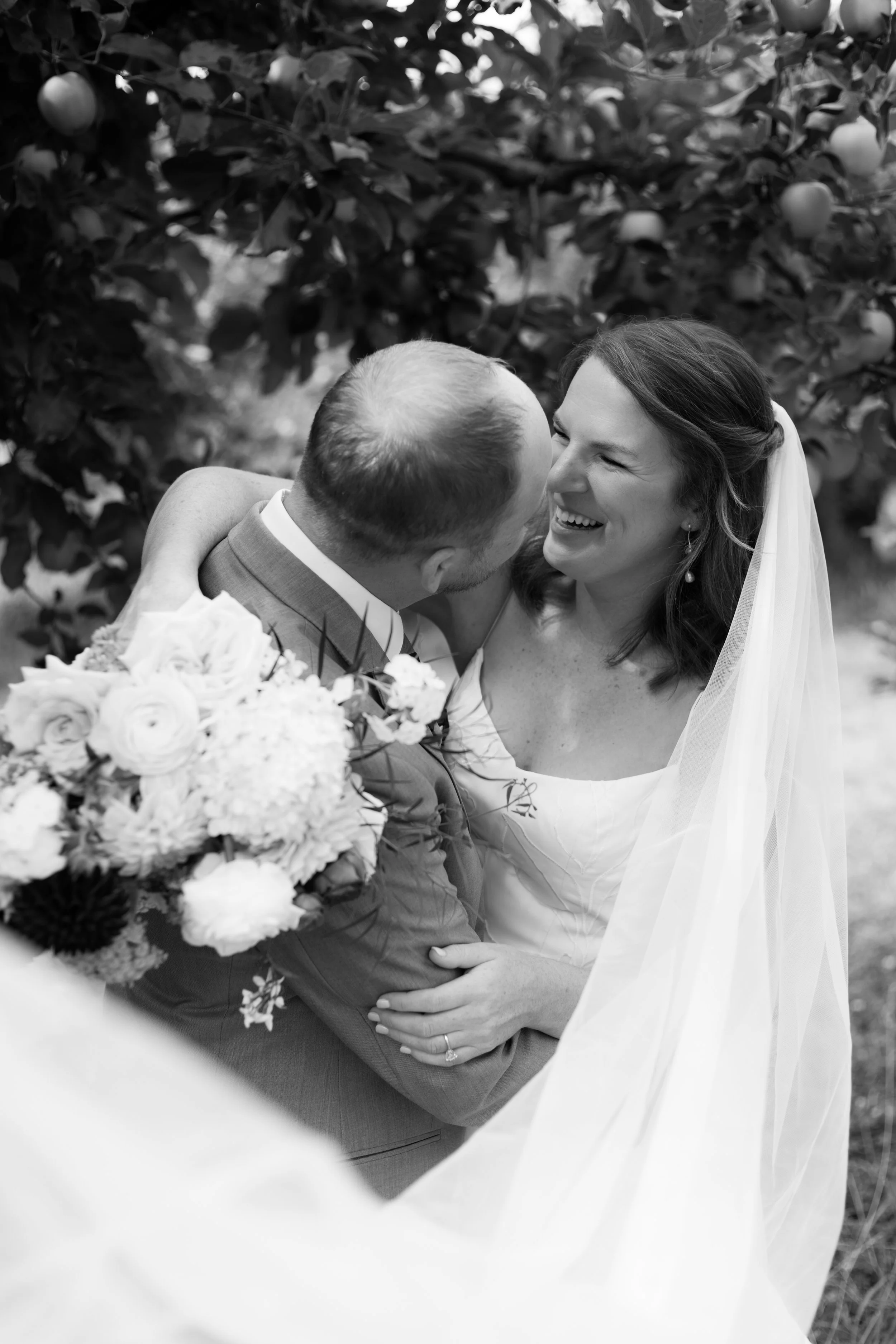 A bride and groom on their wedding day sharing a joyful moment outdoors, surrounded by trees with fruit, with the bride wearing a veil and earrings and holding a bouquet of flowers.