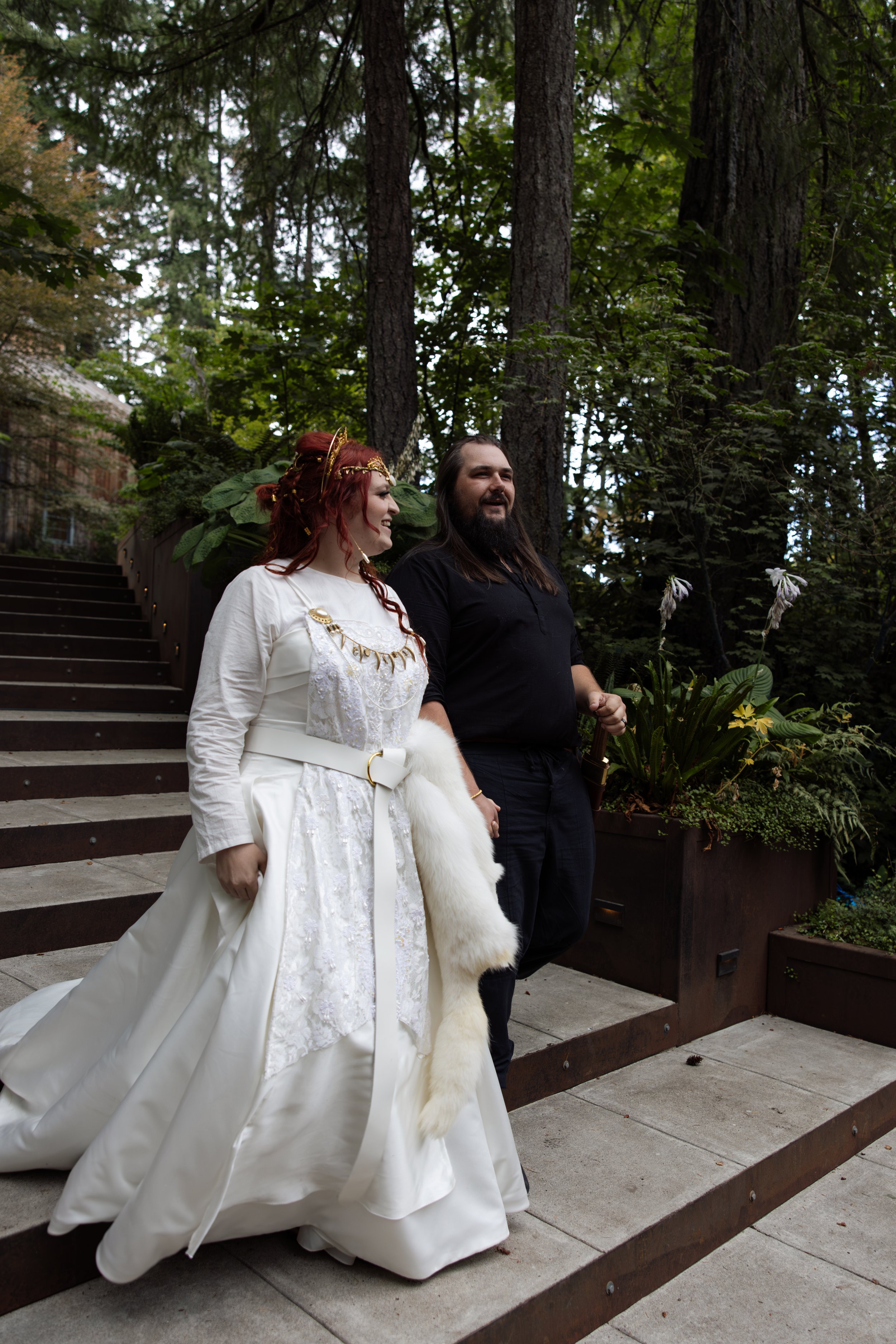 A woman in an elegant white wedding dress with detailed embroidery and a flowing train, holding a white fur stole, walking down outdoor stairs with a man dressed in black, surrounded by tall trees and plants.