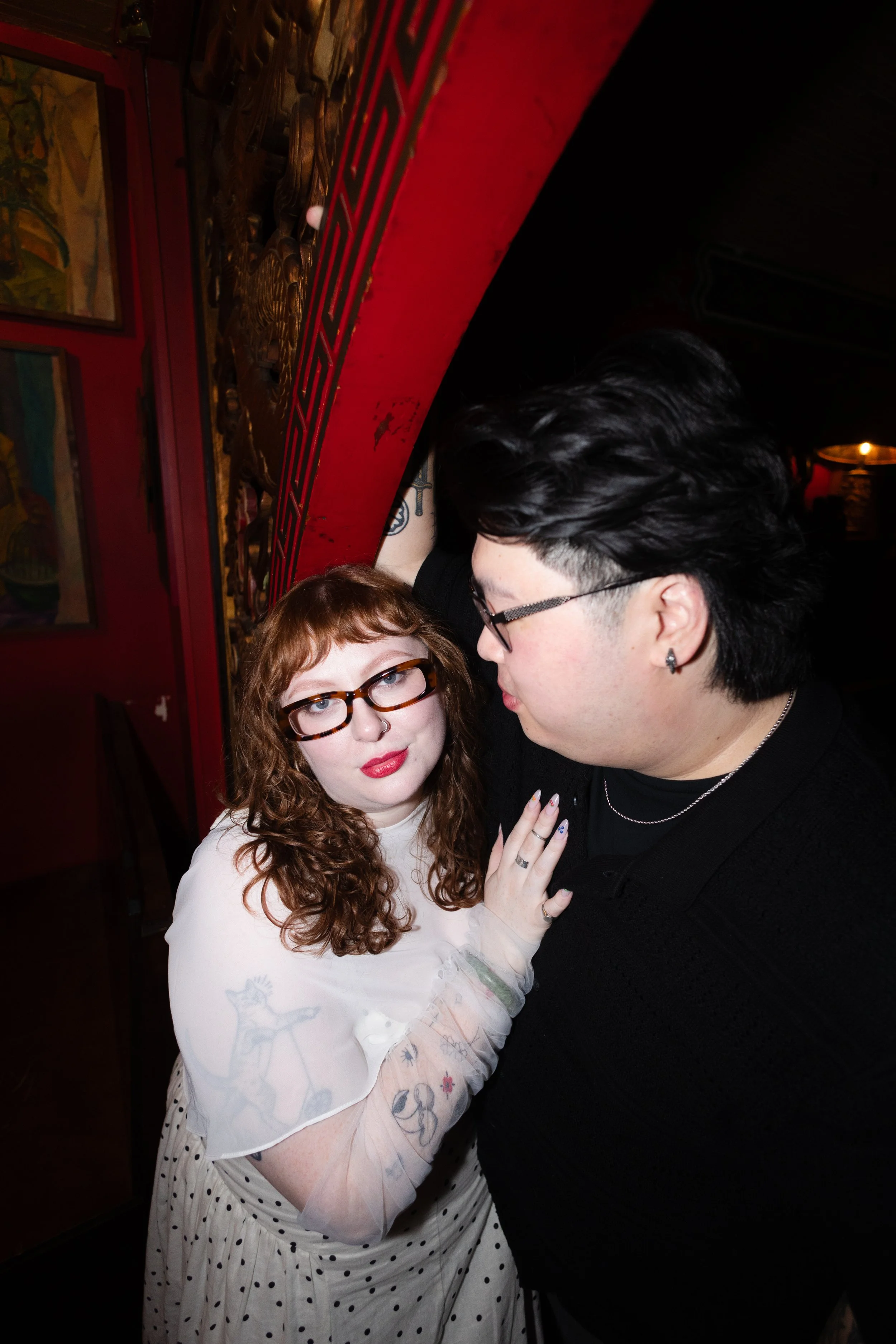 A young woman with red curly hair, glasses, and tattoos on her arms, wearing a white dress with black polka dots, is touching a young man with black hair and glasses. They are close together in a dimly lit room with red walls and framed artwork.
