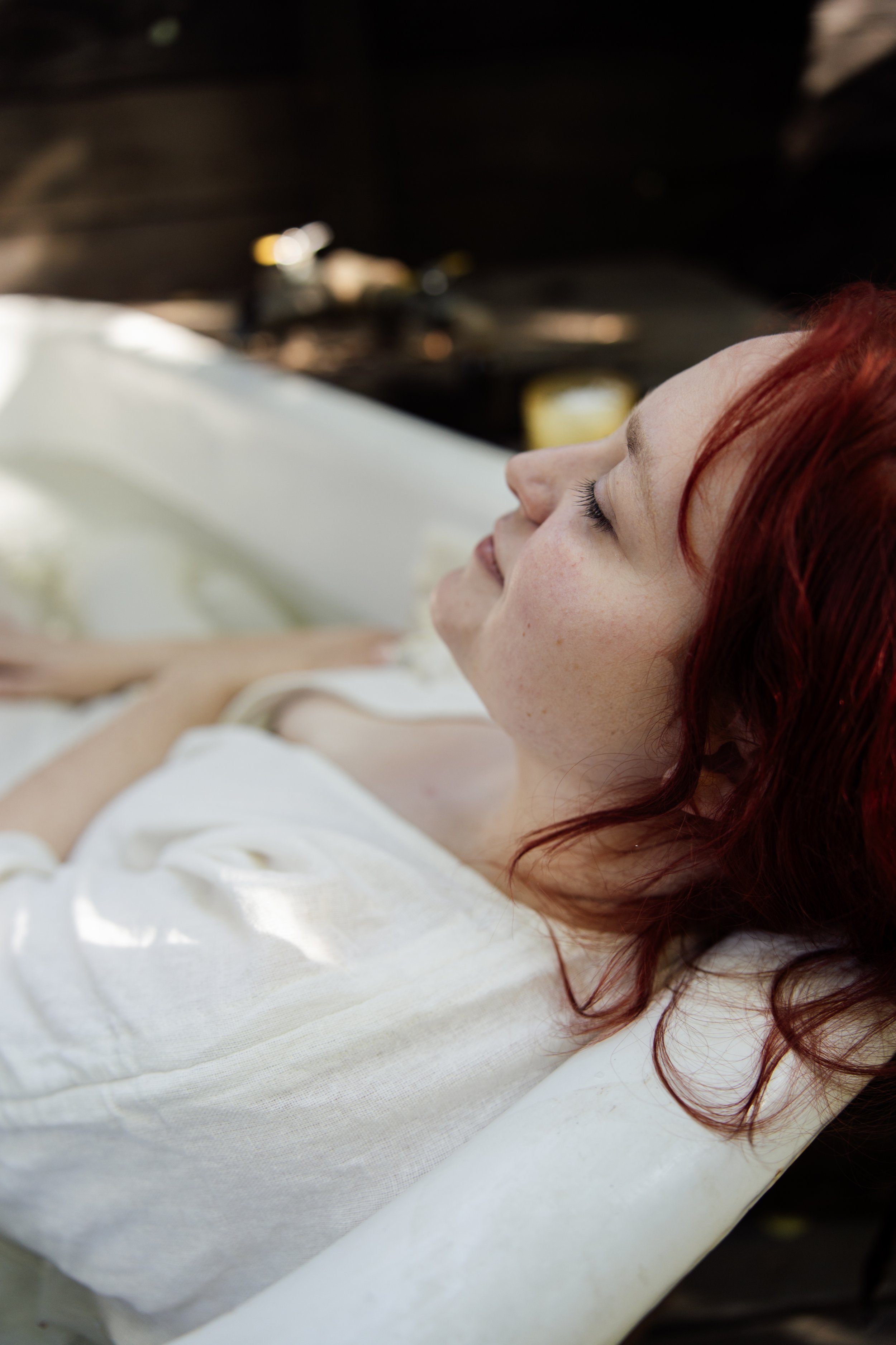 Close-up of a woman with red hair, lying on a white sheet or blanket with her eyes closed.