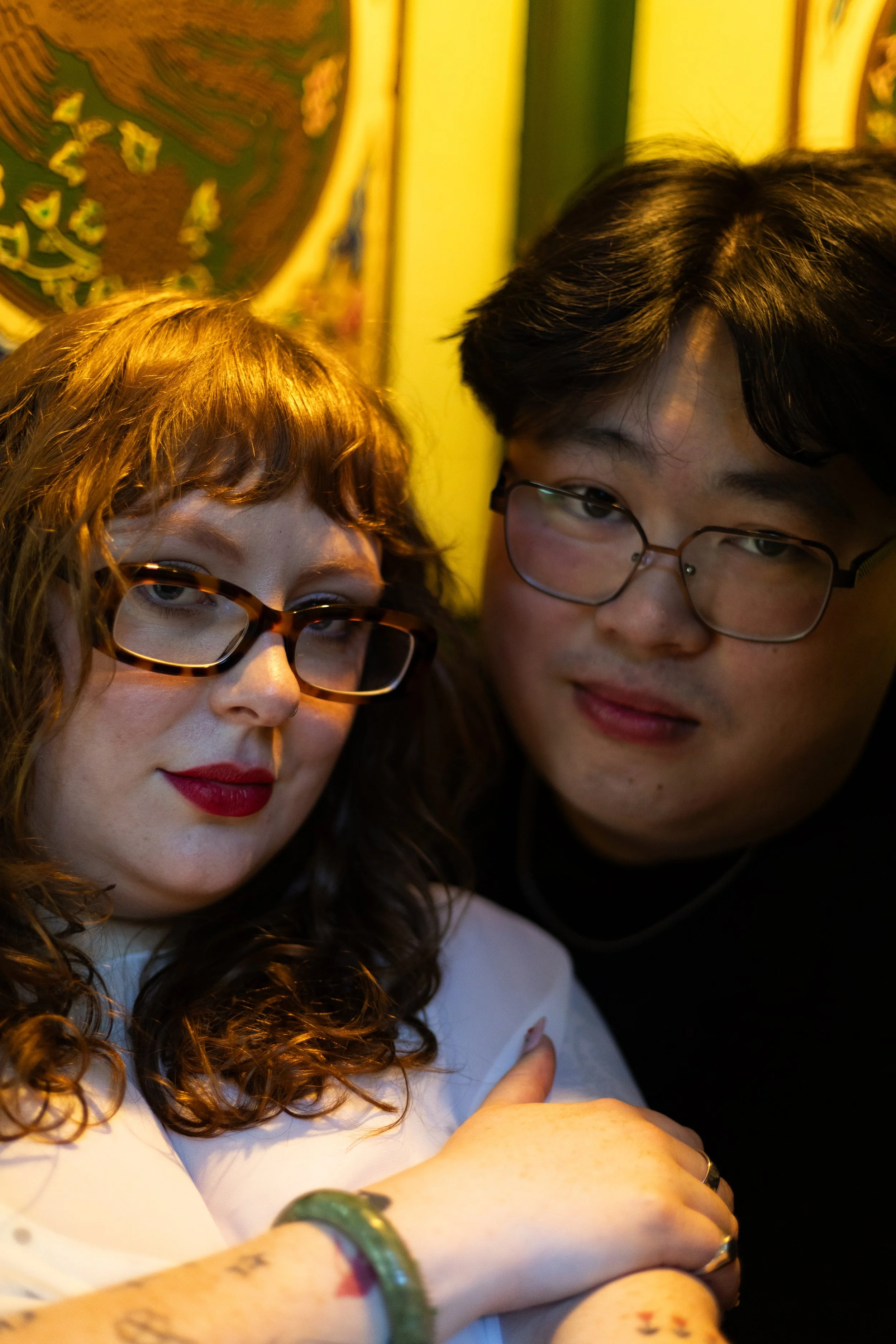Two young women with glasses and wavy hair close together, one with red hair and the other with dark hair, taking a selfie indoors.
