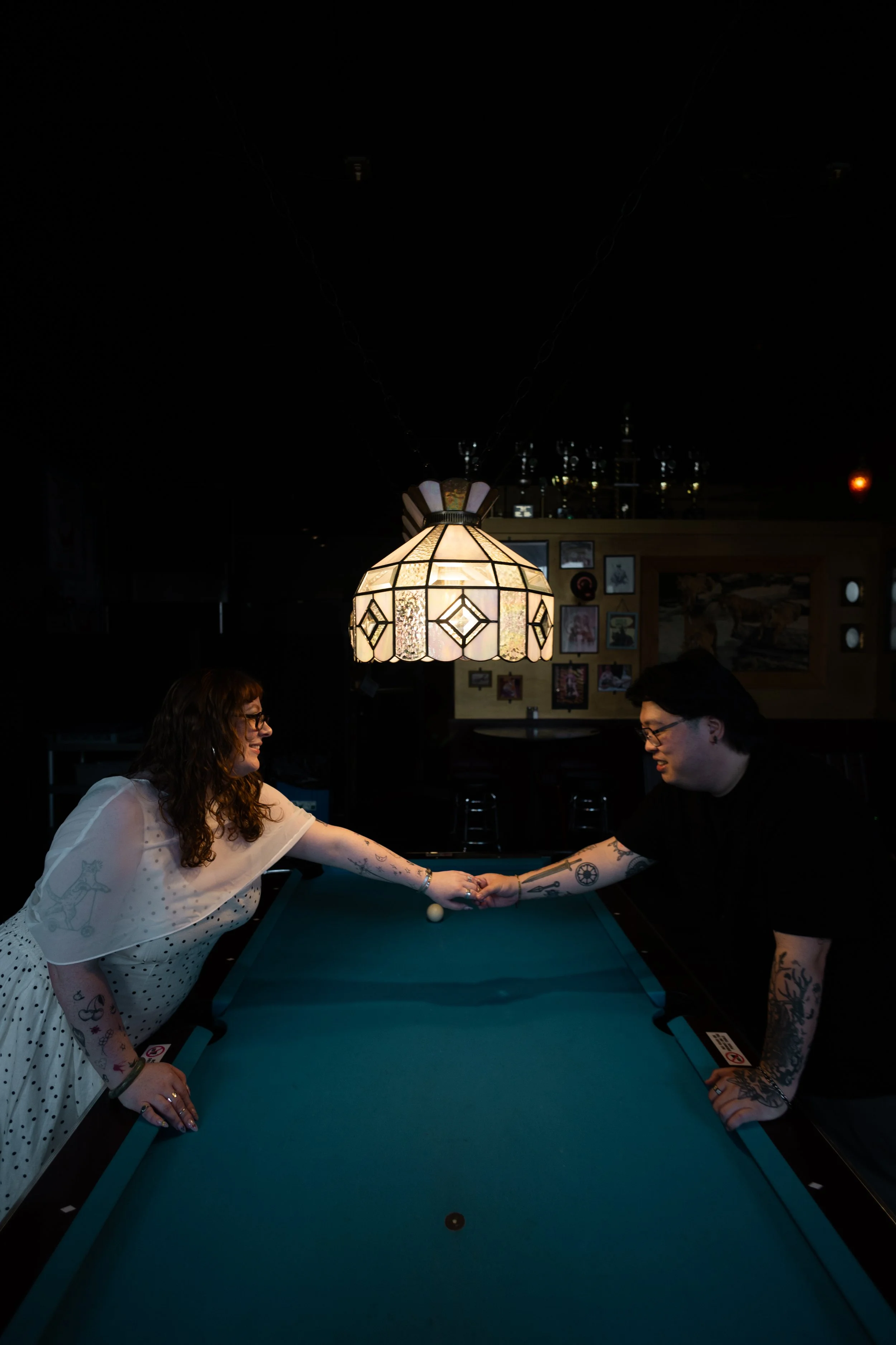 Two people with tattoos, one woman in a white polka-dot dress, and one man in a black shirt, are playing pool together in a dimly-lit room with a hanging stained-glass lamp.
