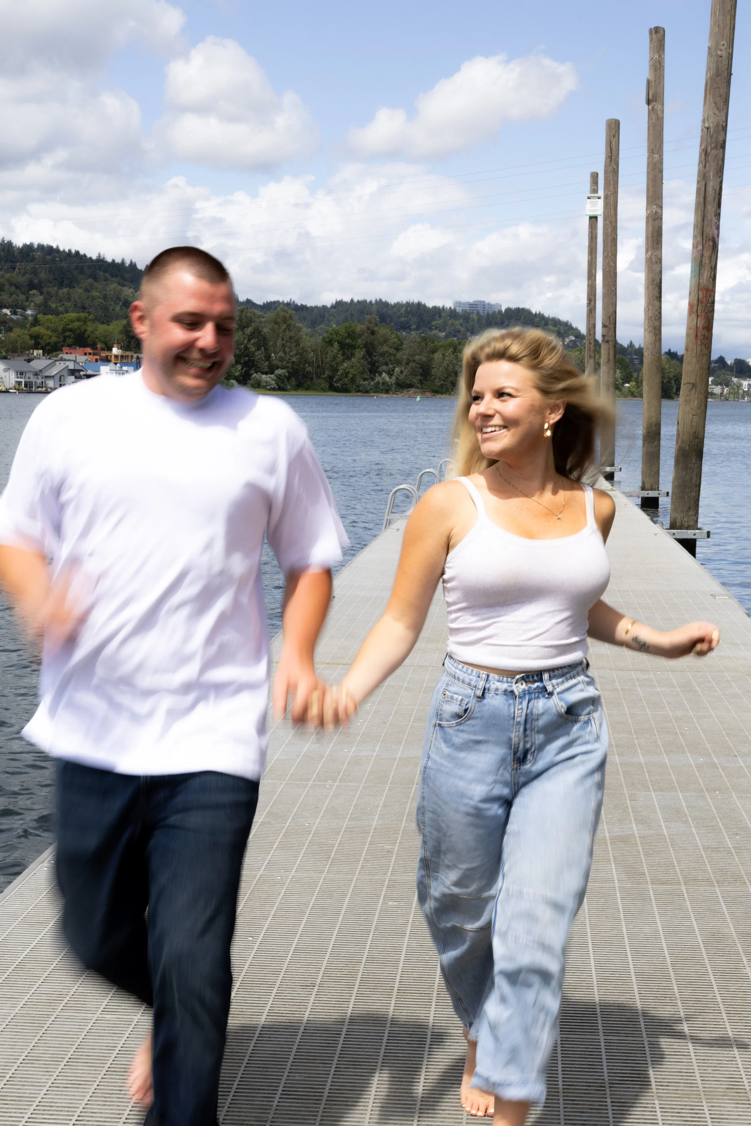 A smiling man and woman holding hands and walking barefoot on a dock by a lake with trees and houses in the background, under cloudy skies.
