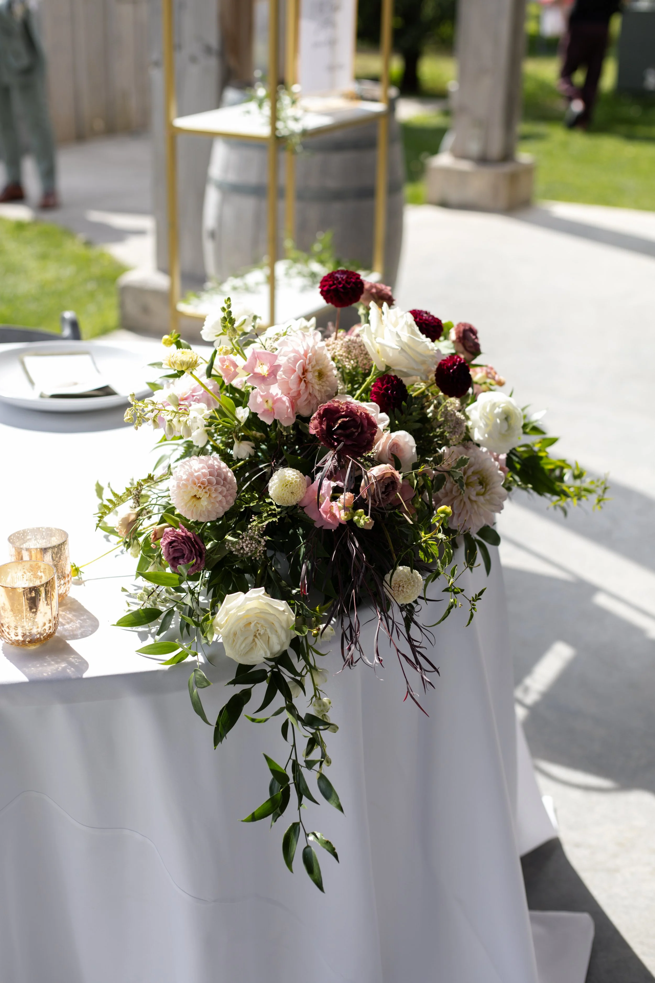 A floral centerpiece with pink, white, and burgundy flowers and greenery on a white tablecloth at an outdoor event.