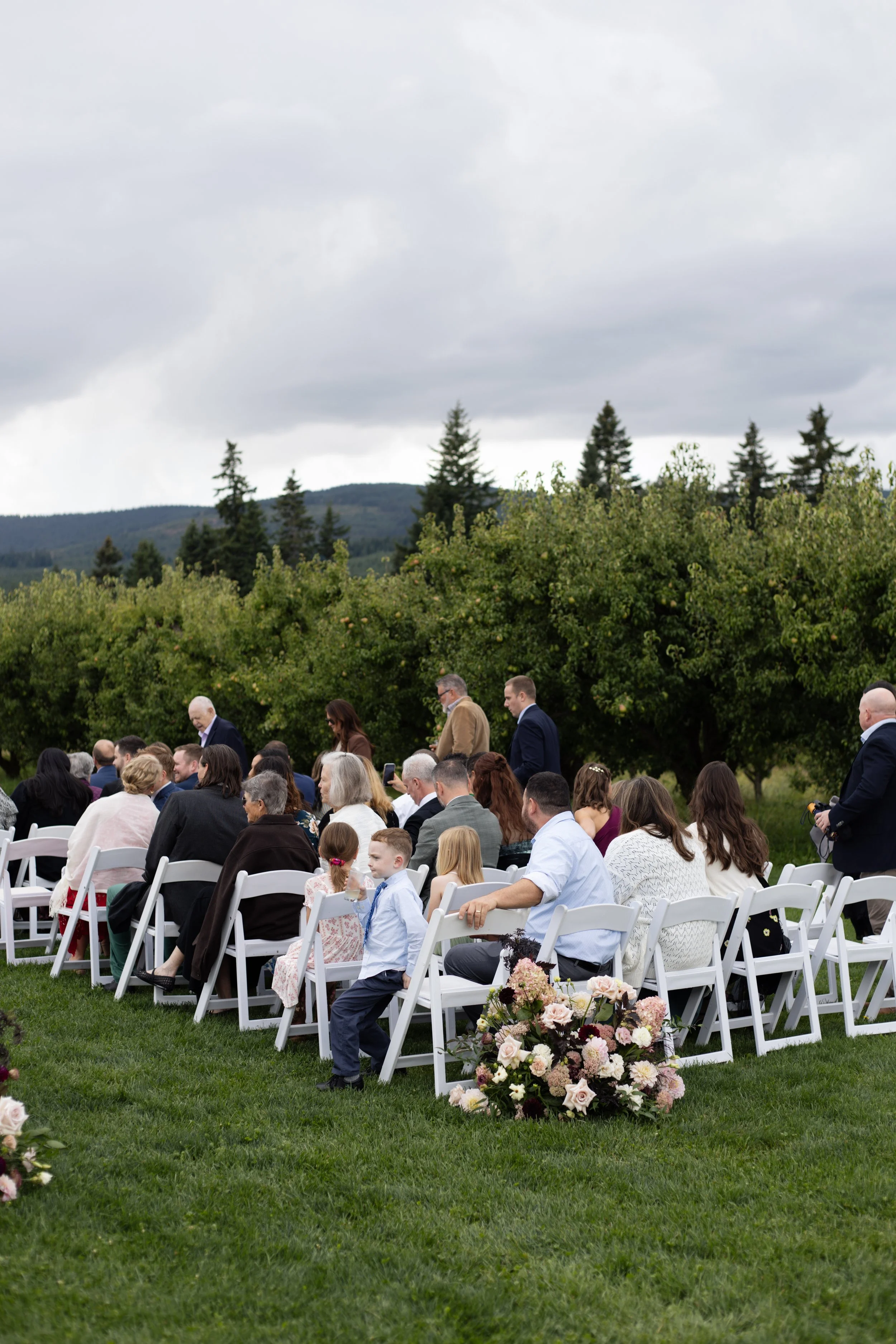 An outdoor wedding ceremony with seated guests, a floral arrangement in the foreground, and a backdrop of green trees and a cloudy sky.