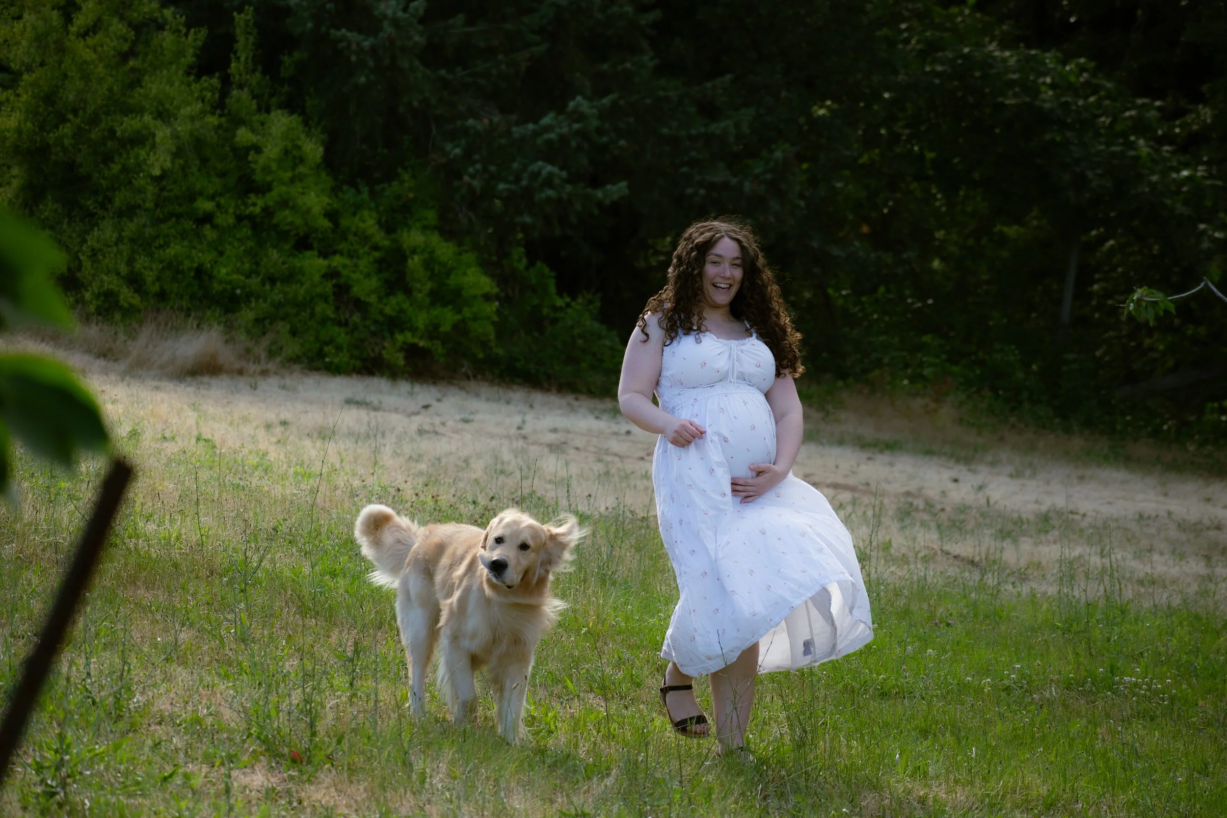 A pregnant woman in a white dress sitting outdoors in a grassy area, smiling and looking down at her belly, with a golden retriever puppy nearby, surrounded by green trees and bushes.