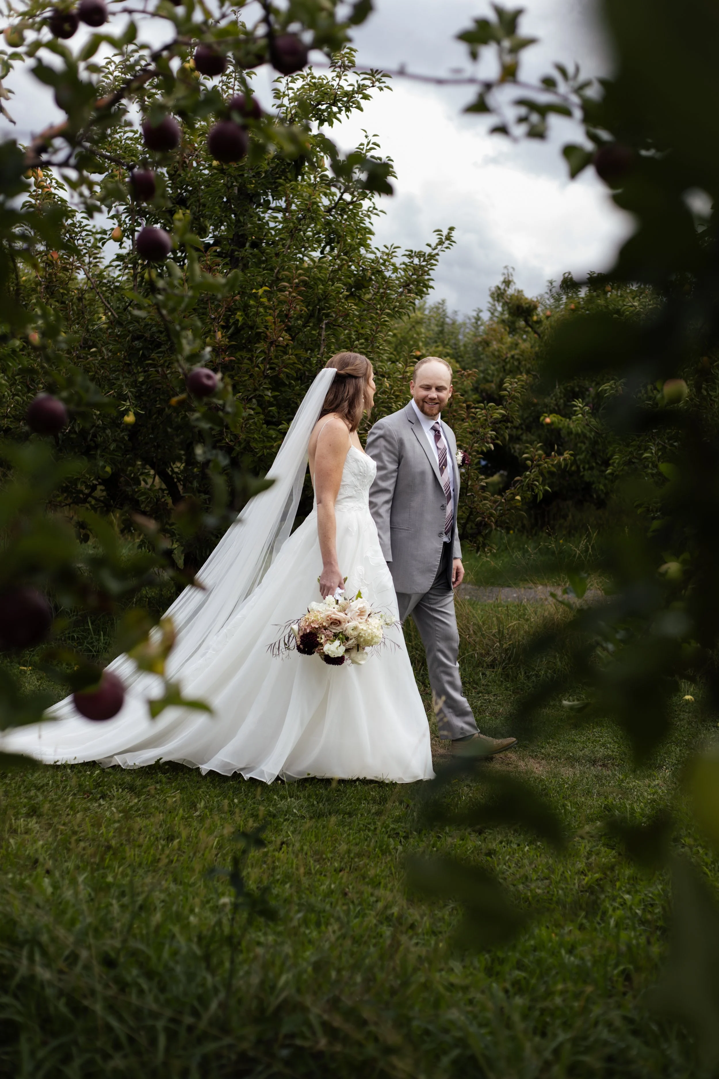 A bride and groom walking in a garden surrounded by apple trees during their wedding photoshoot, with dark clouds in the sky.