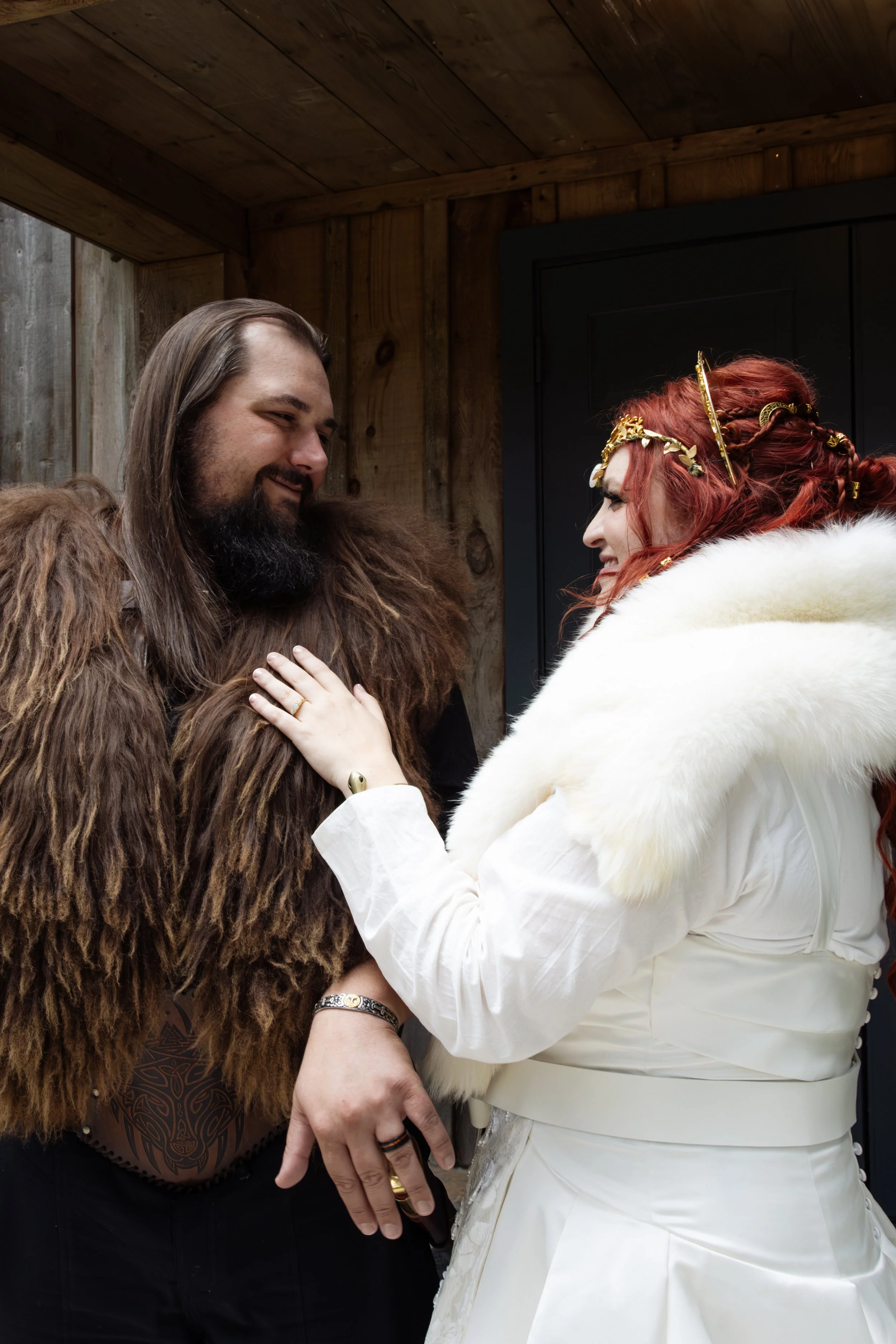 Two people dressed in fantasy costumes sharing an intimate moment indoors. The man, with long brown hair, a beard, and wearing a furry brown cloak, smiles gently. The woman, with elaborate red hair decorated with golden accessories, is wearing white 