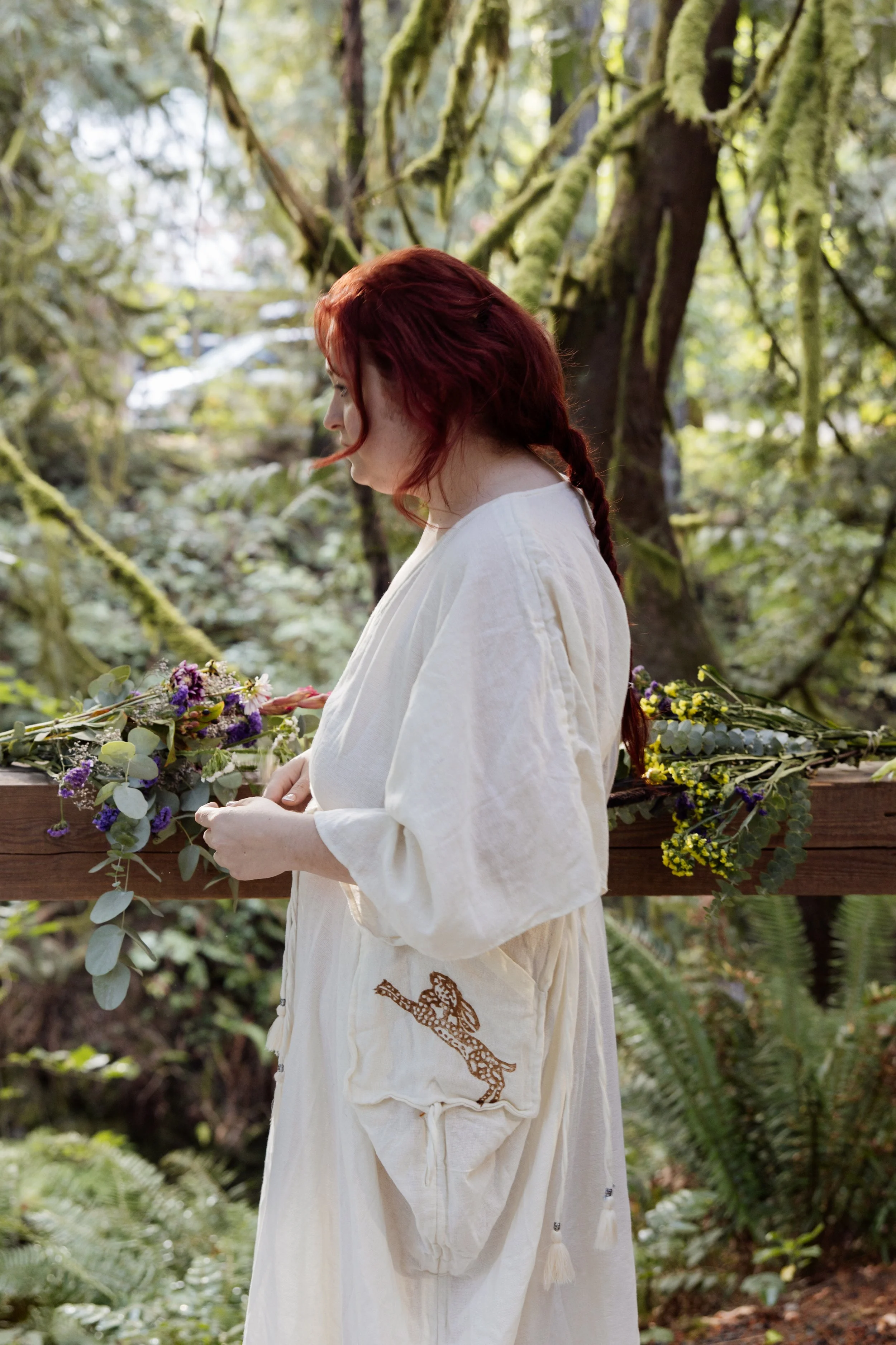 A woman with red hair styled in a braid, wearing a white dress with an embroidered animal on the sleeve, stands outdoors in a lush, green forested area, holding a bouquet of flowers and leaves.