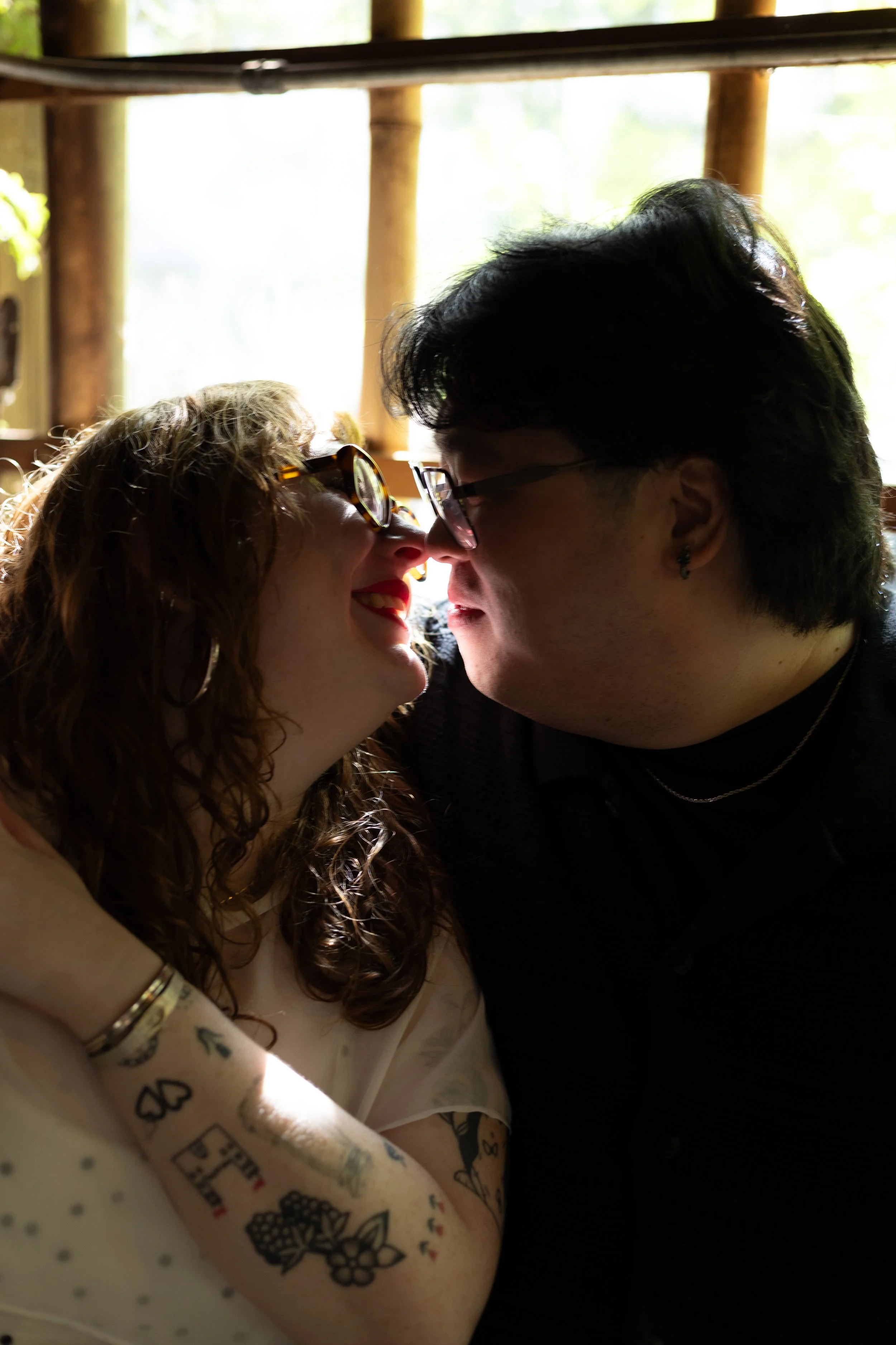 A woman and a man are close, touching noses, smiling, and looking at each other, with the woman wearing sunglasses and having tattoos on her arm, and the man wearing glasses.