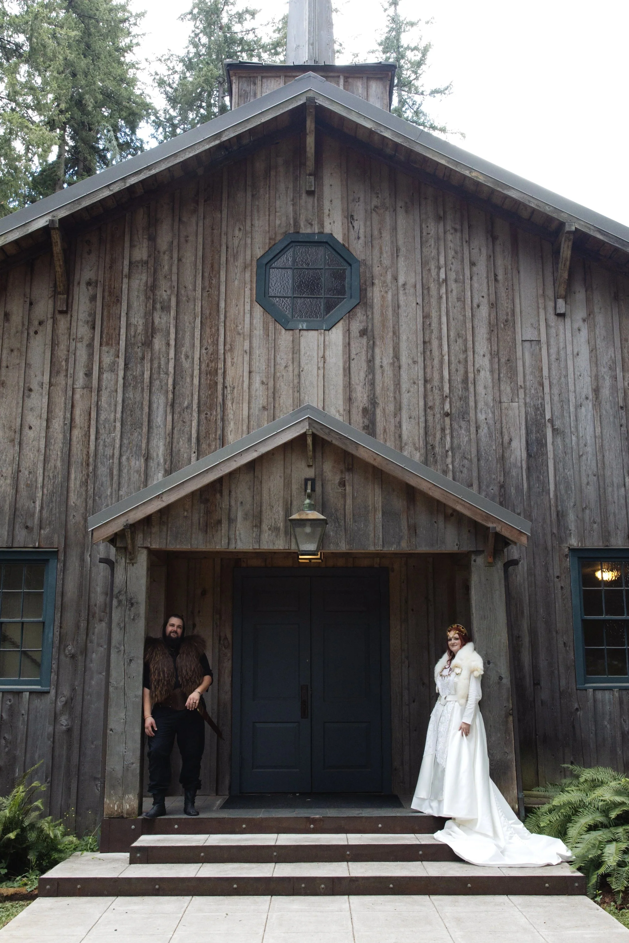 A man with a beard wearing a fur vest and black pants, and a woman in a white wedding dress with a fur shawl, standing in front of a rustic wooden building with steps and trees in the background.
