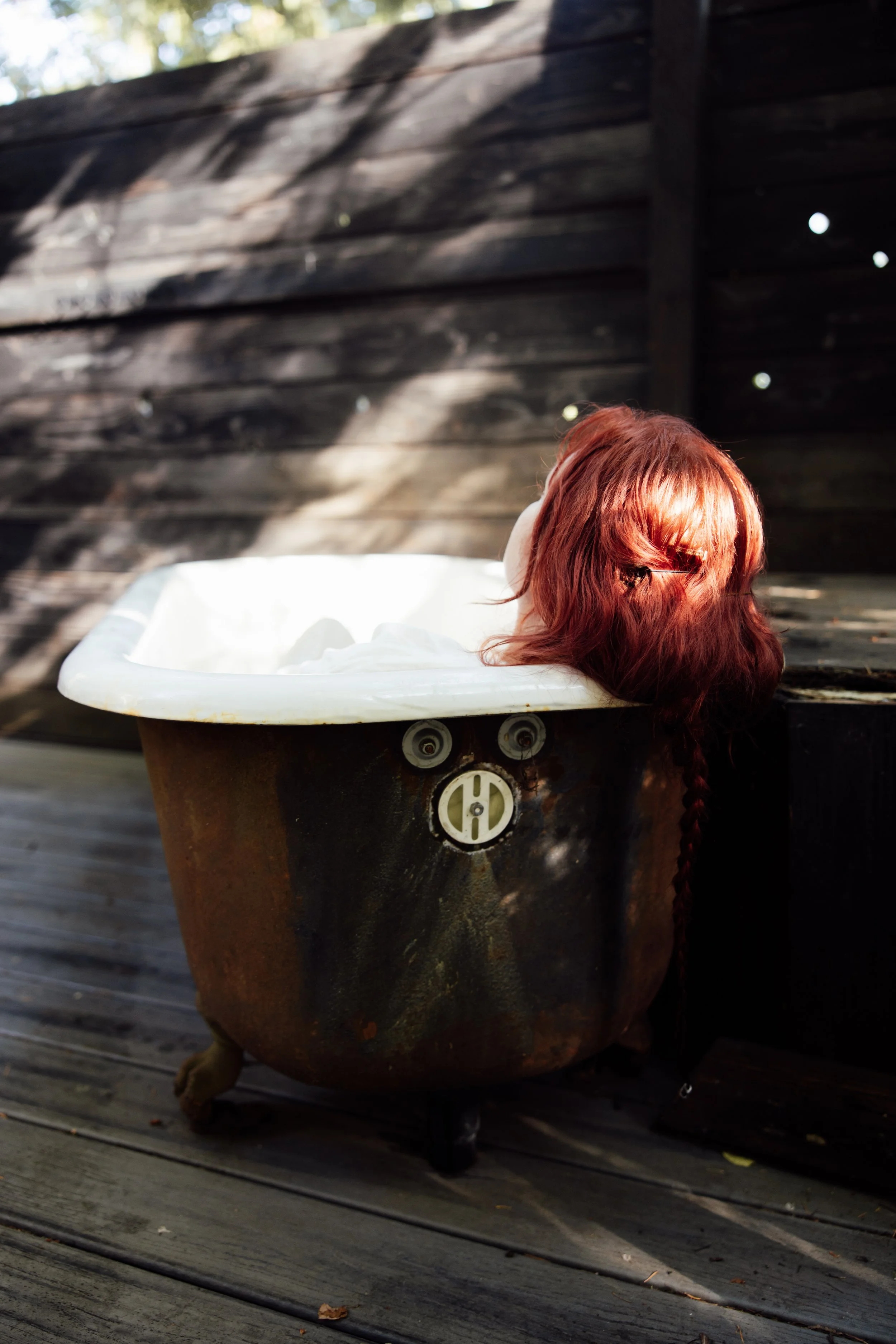 A person with red hair is reclining in a vintage bathtub outdoors on a wooden deck, with a rustic dark wooden fence in the background and sunlight filtering through trees.