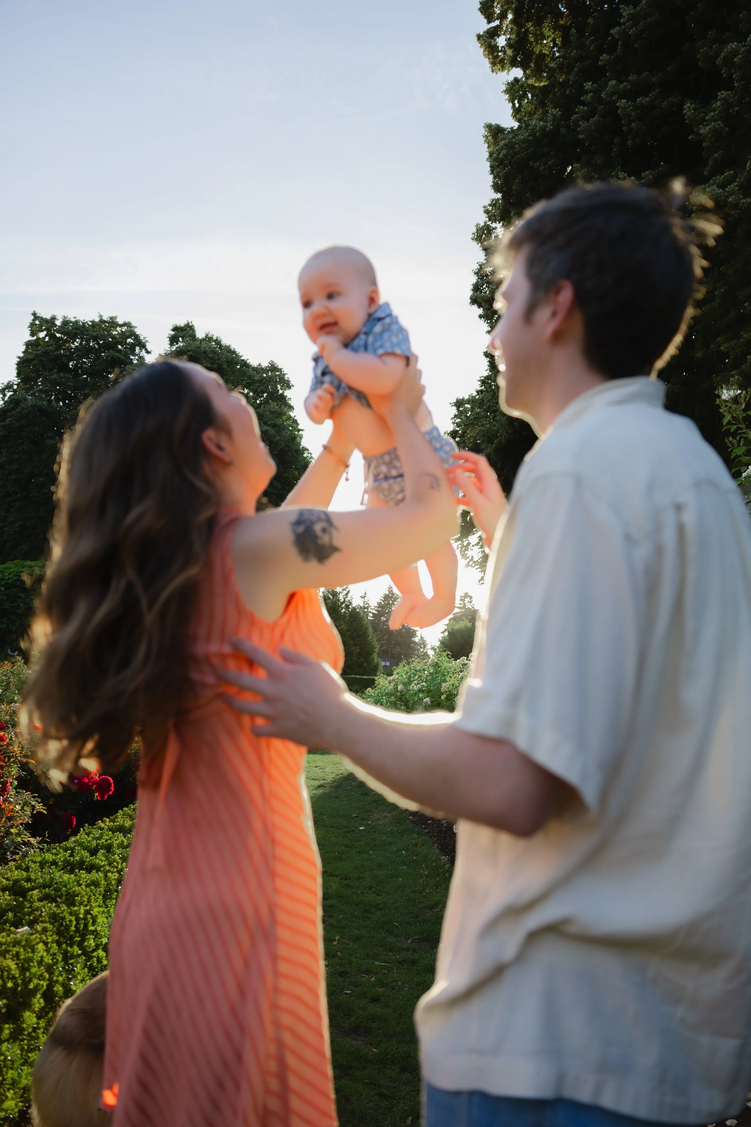 A woman lifting a baby in the air outdoors with a man standing nearby, trees and a clear sky in the background, during sunset.