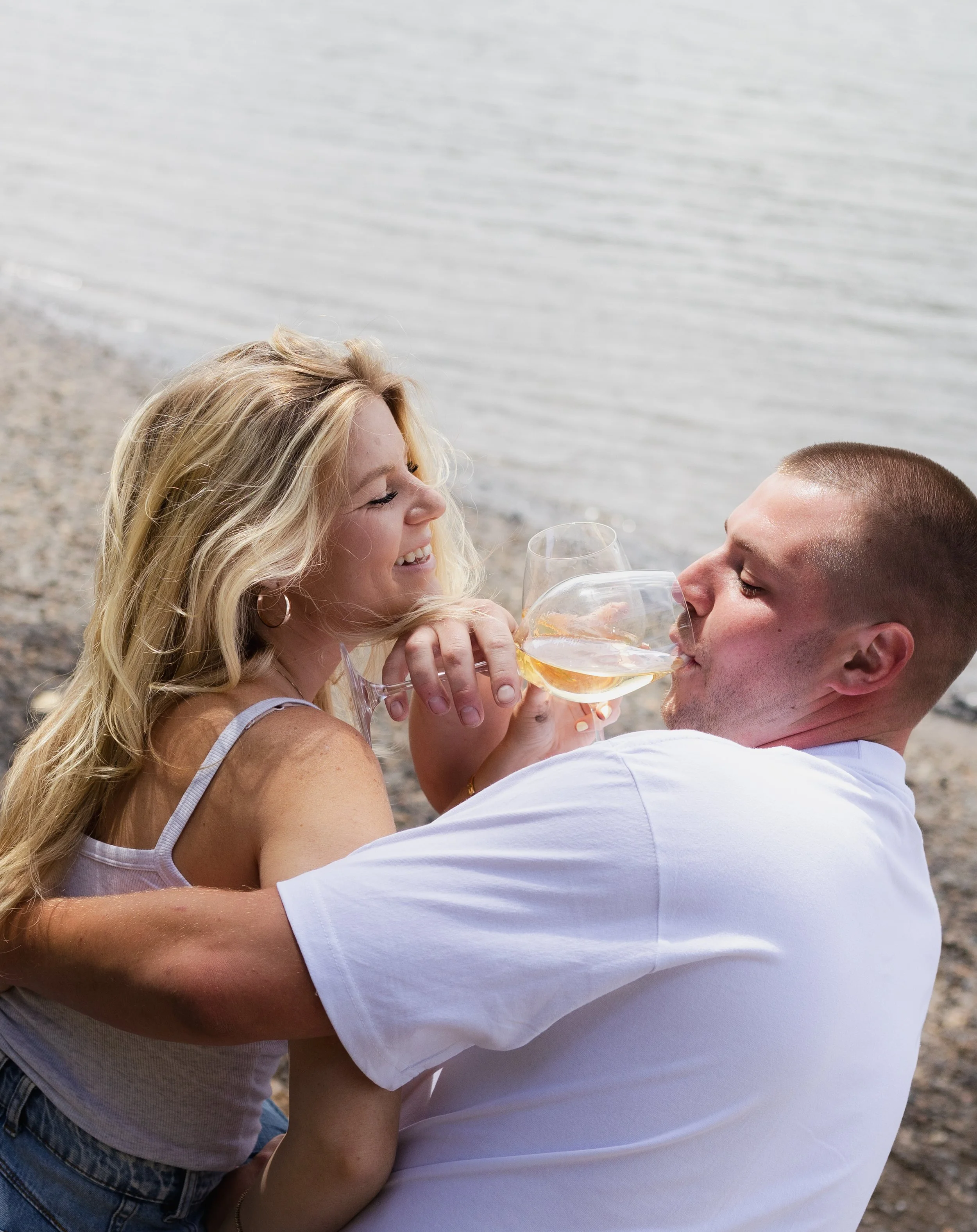 A woman and a man smiling and clinking wine glasses together by the water on a beach.