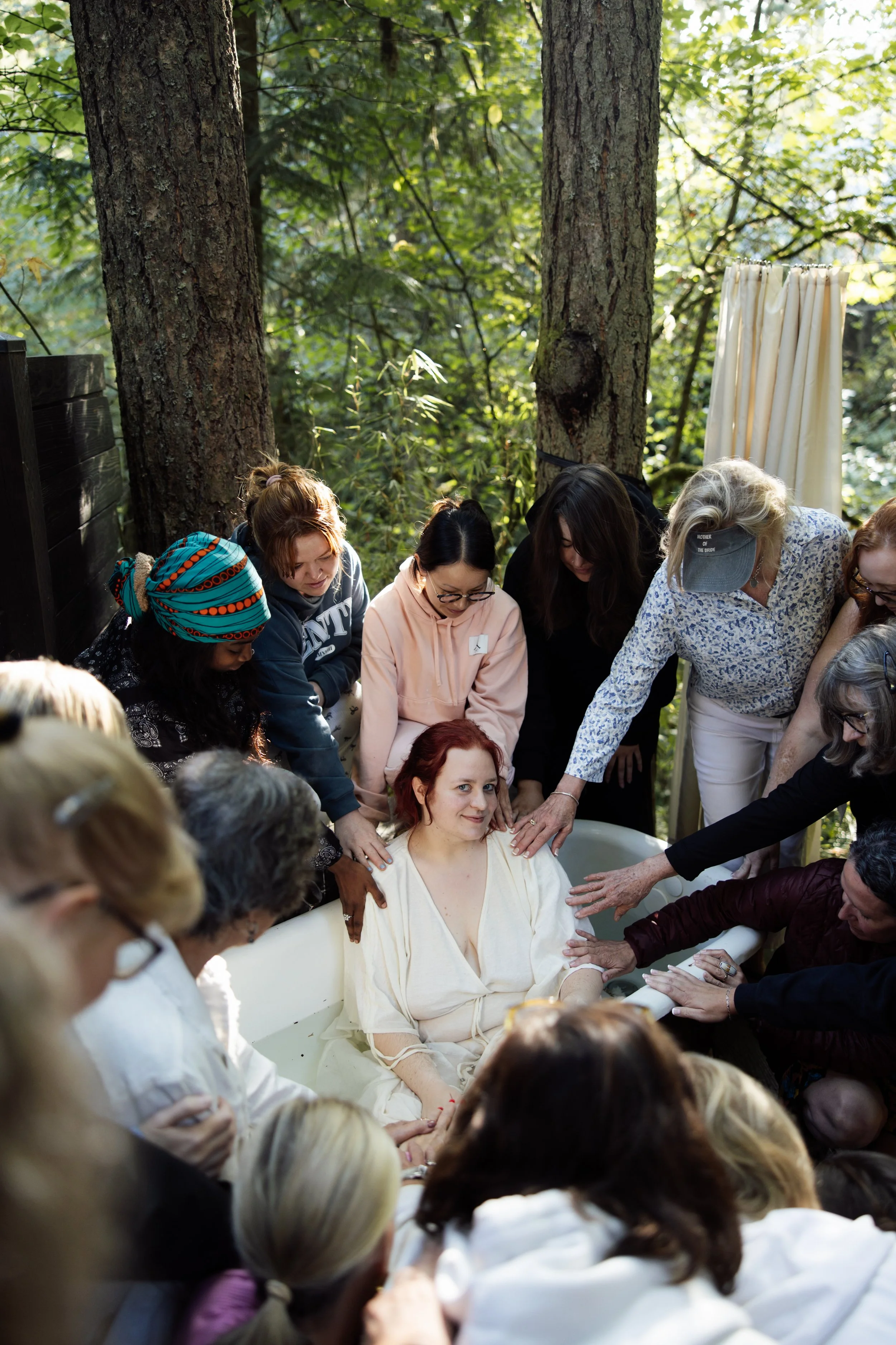 A group of people gathered outdoors in a wooded area, surrounding a woman in a light-colored dress sitting in a bathtub, as they participate in a communal or ceremonial activity.