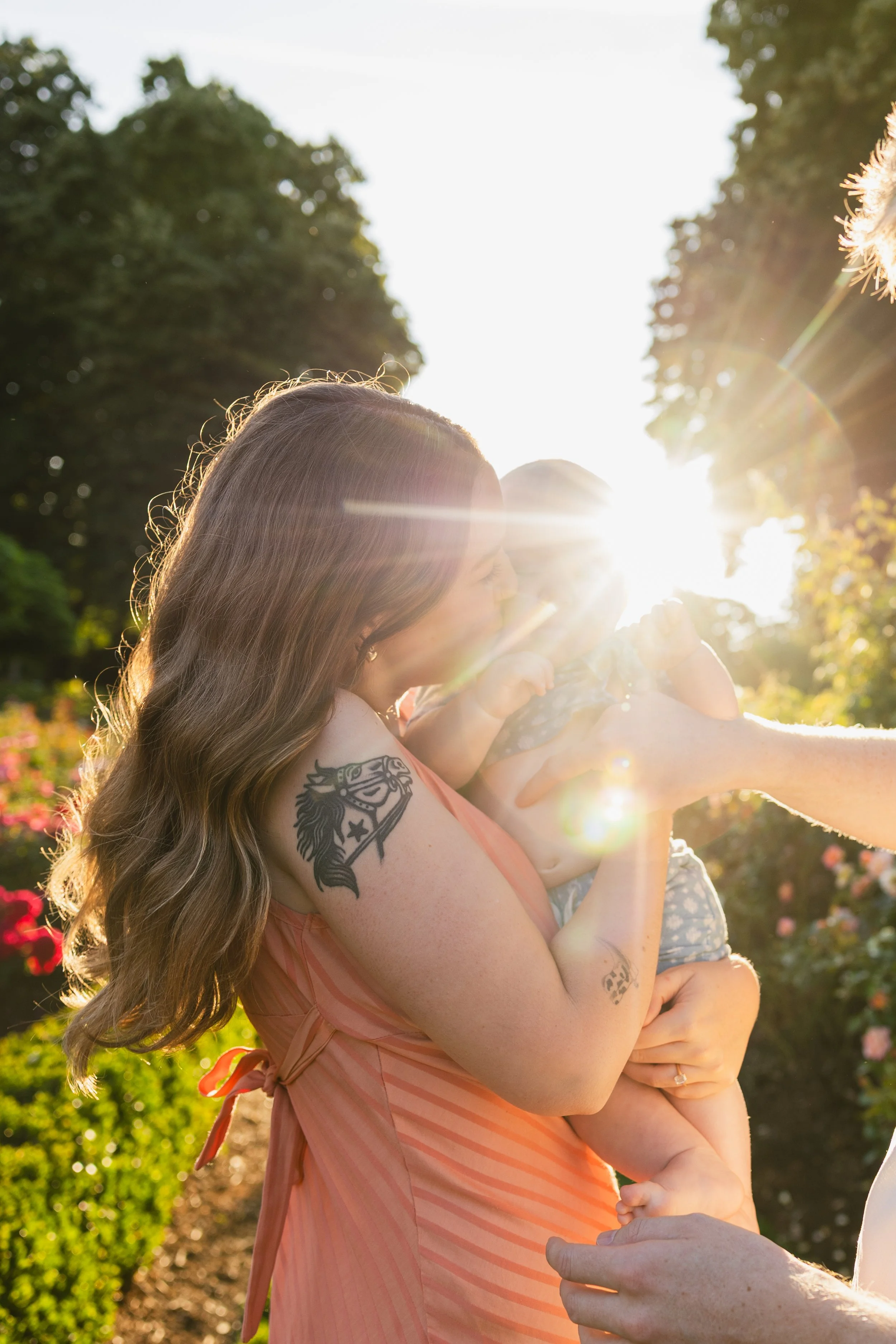 A woman with long wavy hair and tattoos on her arm holding a baby outdoors in a park at sunset, with sunlight shining through the trees.