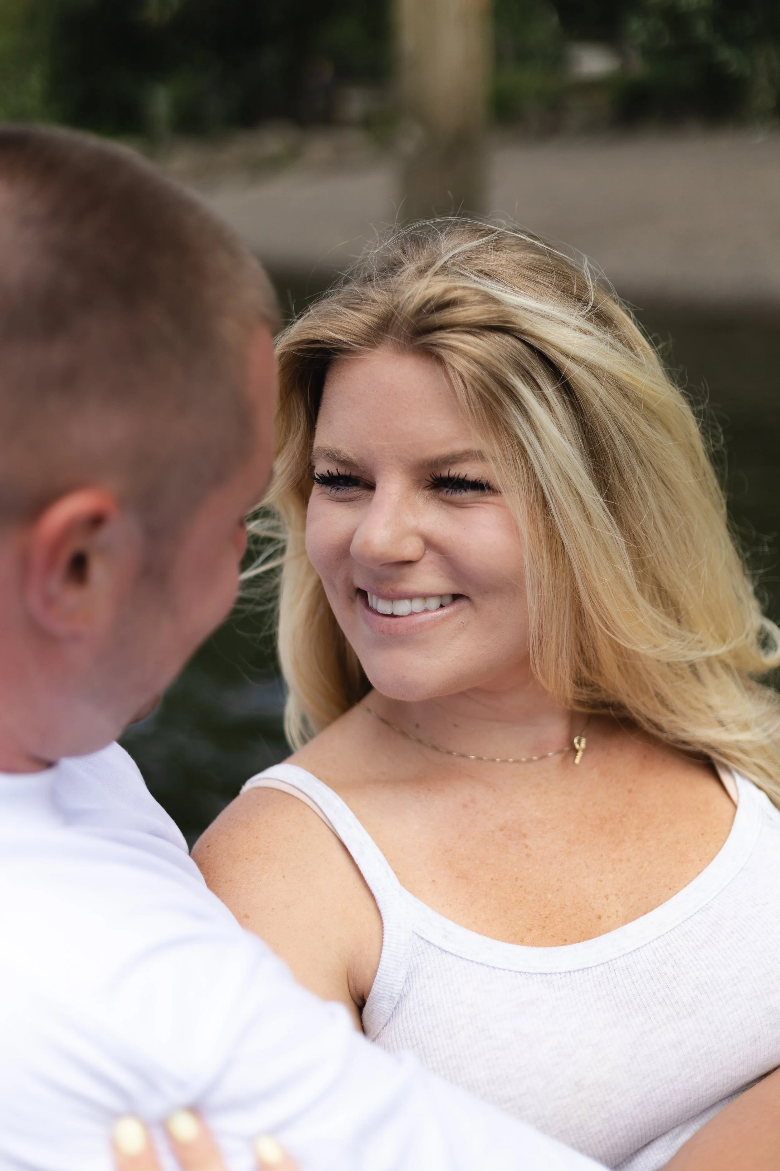 A smiling woman with blonde hair in a white tank top looking at a man in a white shirt outdoors.