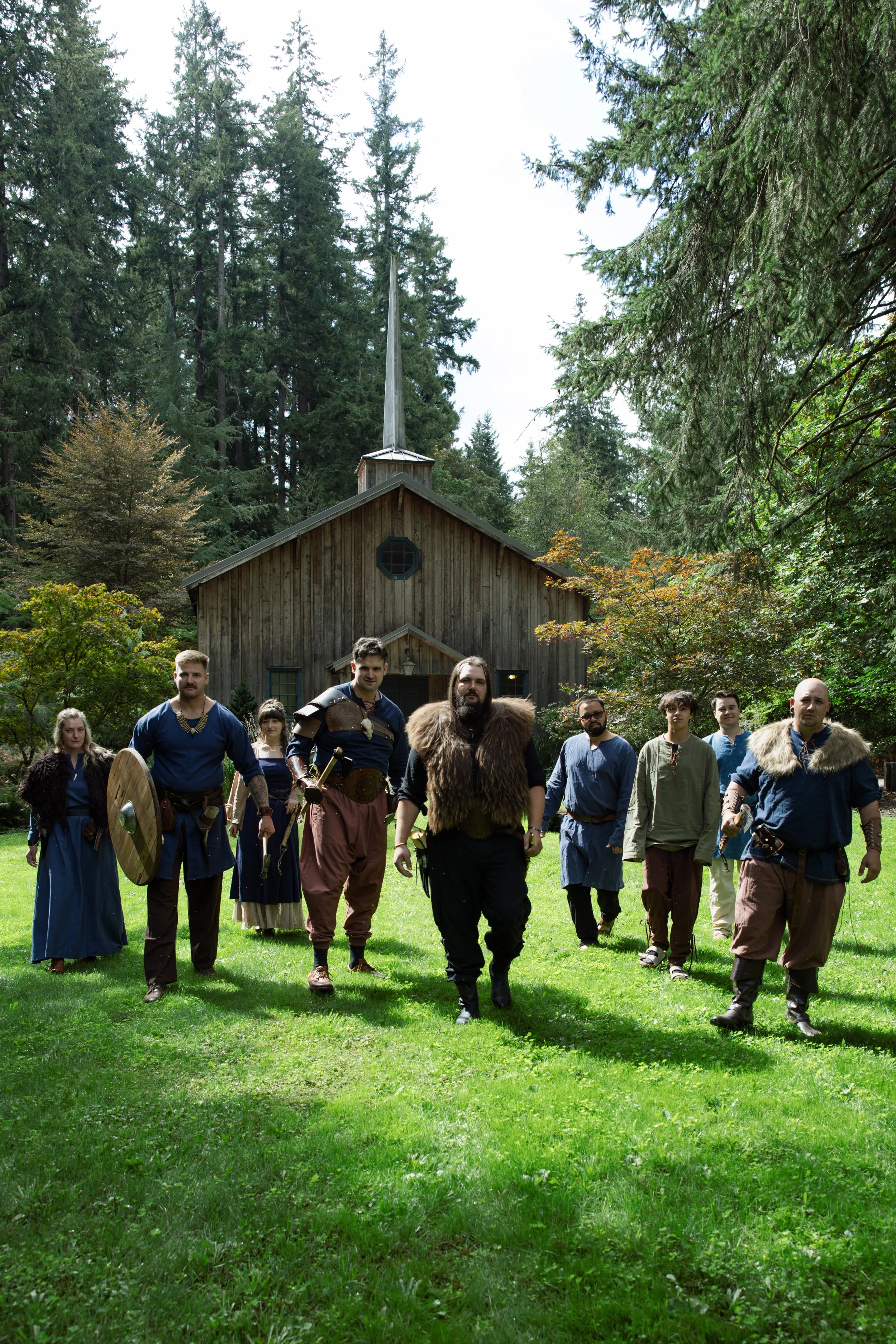 A group of people dressed in medieval or fantasy costumes walking on a grassy area in front of a wooden church, surrounded by tall green trees.