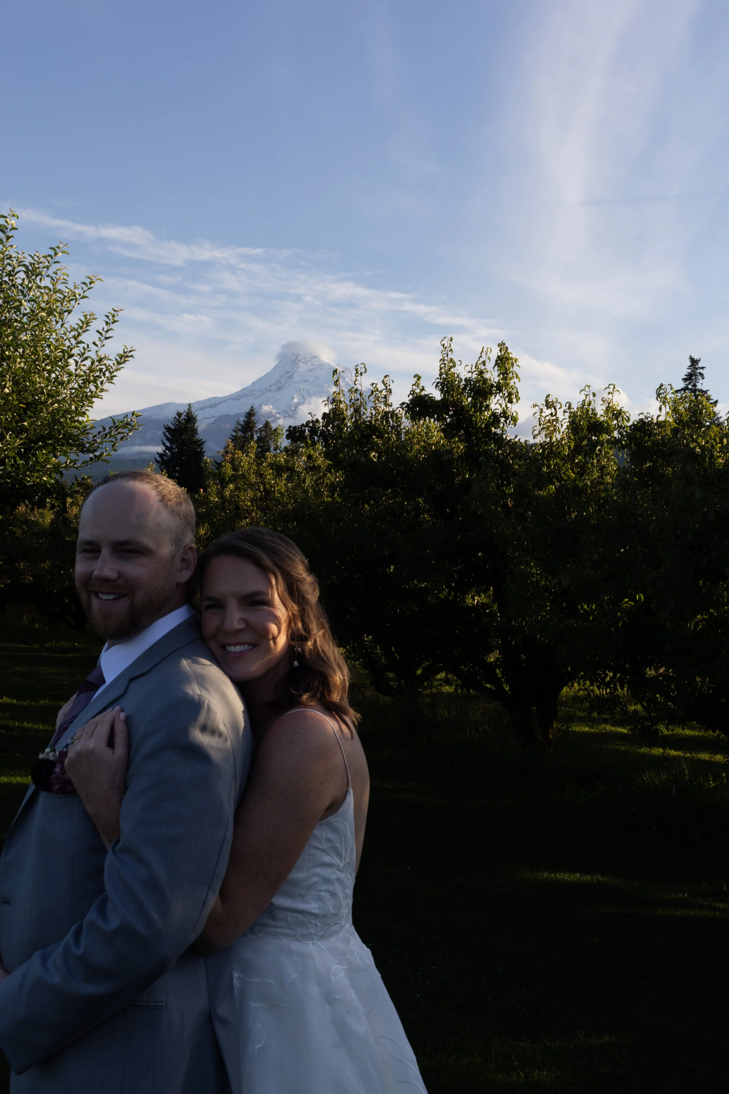 A smiling couple stands together outdoors in front of a lush green landscape with trees, with a snow-capped mountain in the background under a partly cloudy sky.