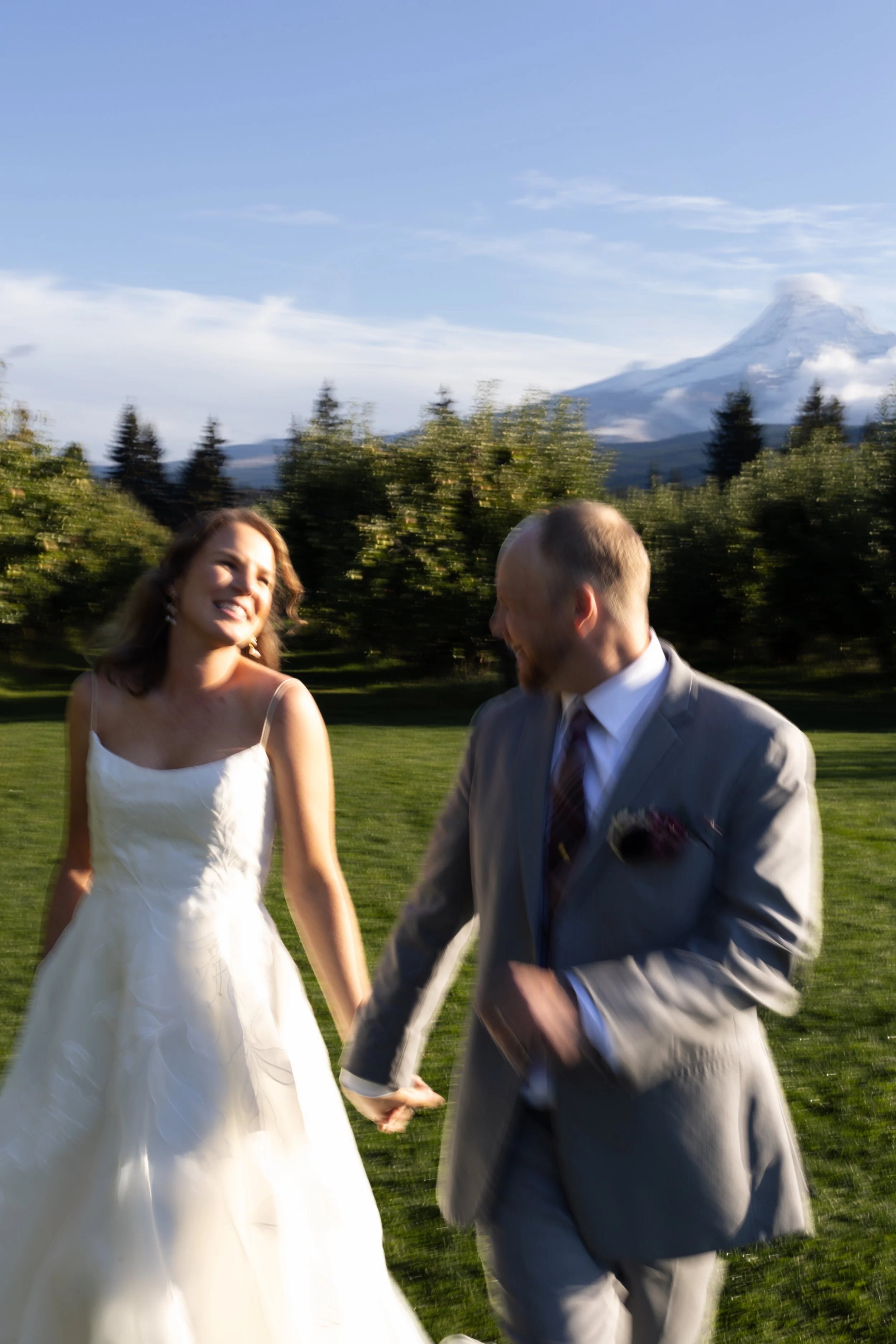 A smiling bride and groom holding hands, walking outdoors on grass with a blurred background of trees and a mountain with snow, under a clear blue sky.