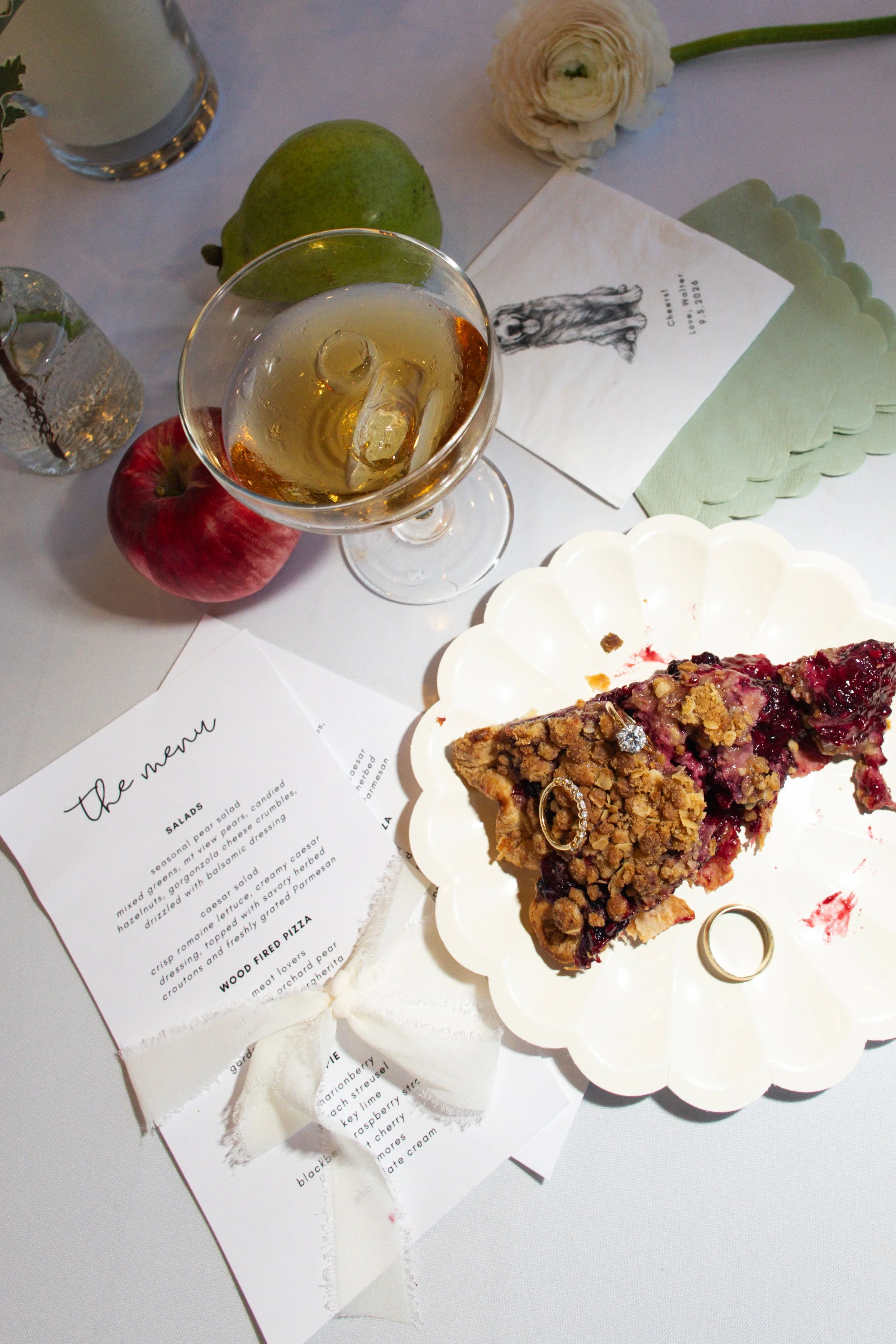 A table setting with a slice of berry crumble pie on a white scalloped plate, a glass of white wine with ice, apple and pear, a white flower, a glass bottle of water, and a menu sheet with food descriptions.