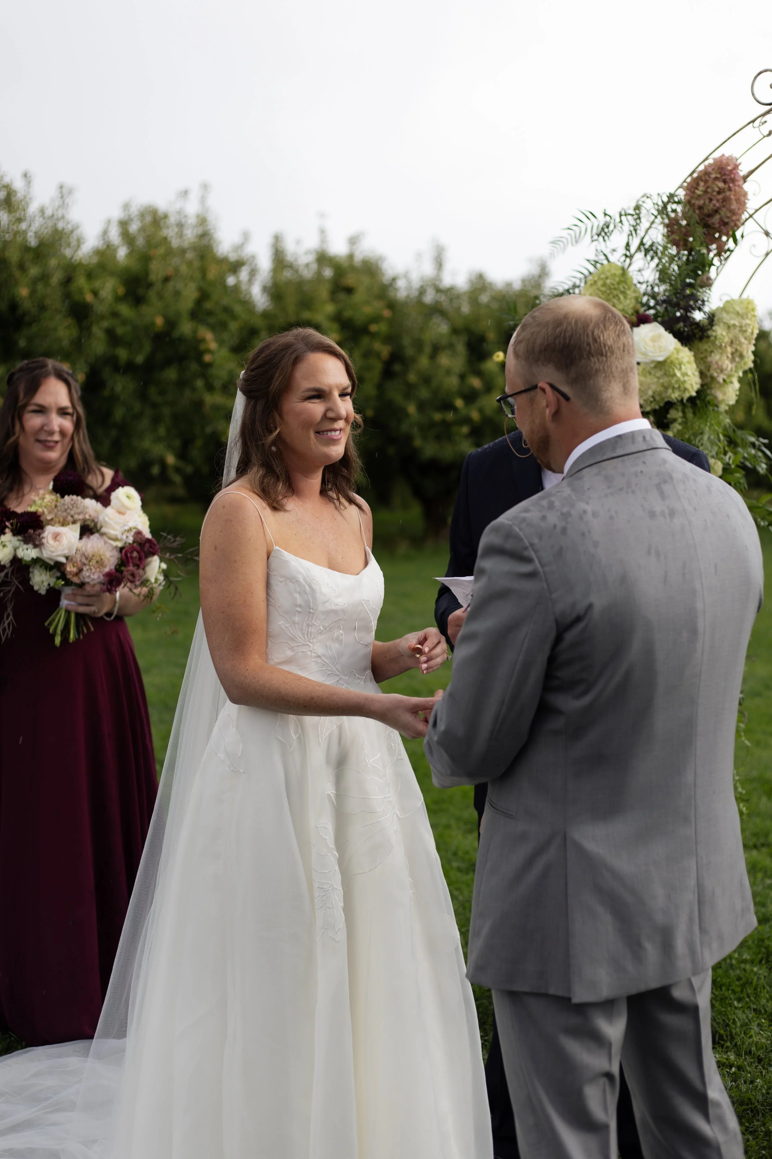 A wedding ceremony outdoors with a bride in a white dress and a groom in a gray suit holding hands, exchanging vows, with a bridesmaid holding a bouquet of flowers in the background.