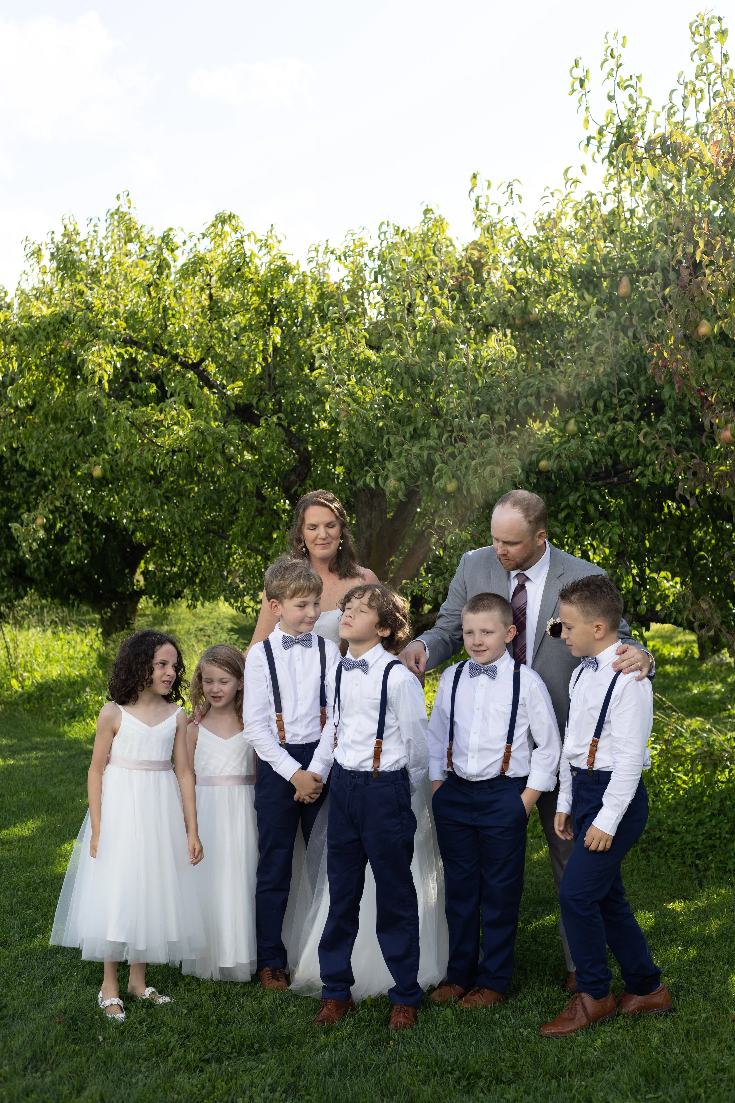 A family gathered outdoors in a garden under a large apple tree, with children dressed in formal attire, including suits, bow ties, and white dresses. They appear to be attending a celebration or special event.