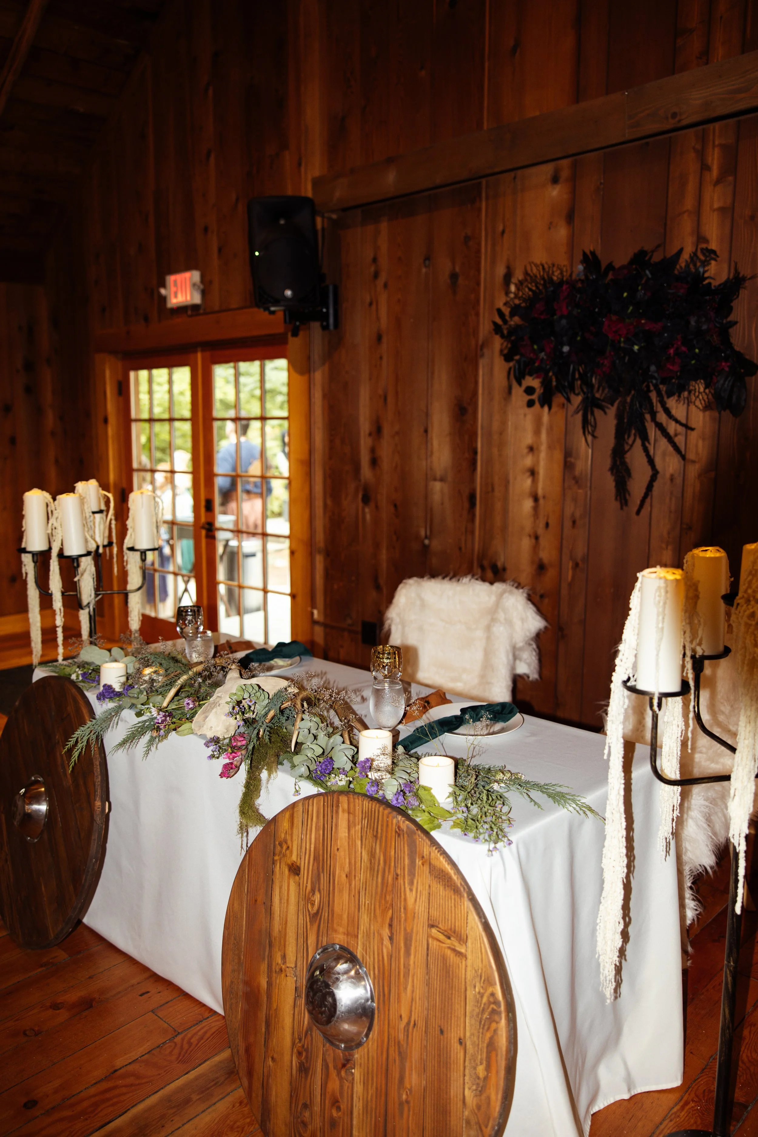 A decorated indoor dining table with a white tablecloth, floral centerpiece, and candles, set in a wooden-paneled room with a door and window in the background.