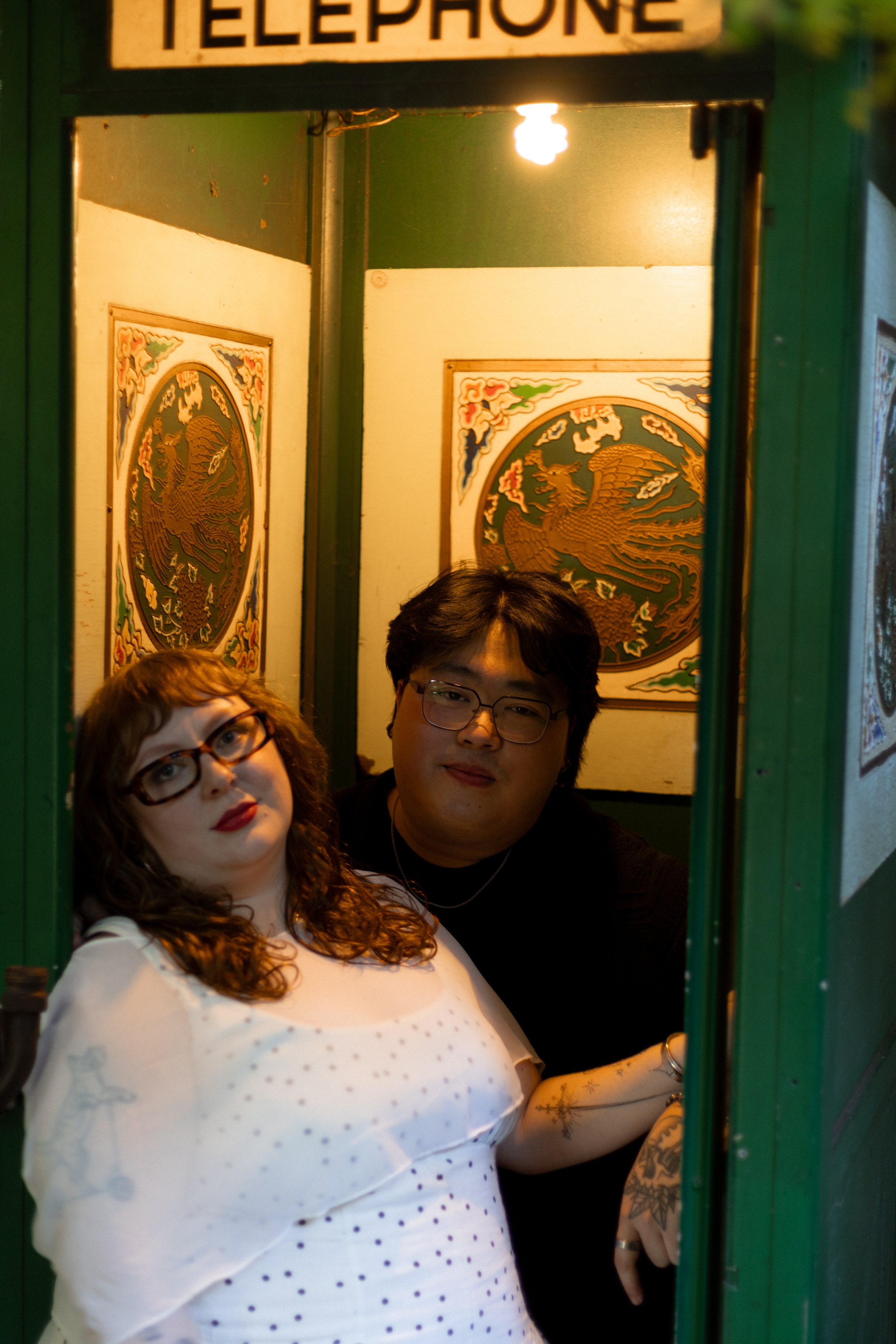 Two people taking a selfie inside an old-fashioned telephone booth decorated with Asian-themed artwork, with a woman with glasses, curly hair, and a white polka-dot shirt in the foreground, and a man with glasses and dark hair behind her.