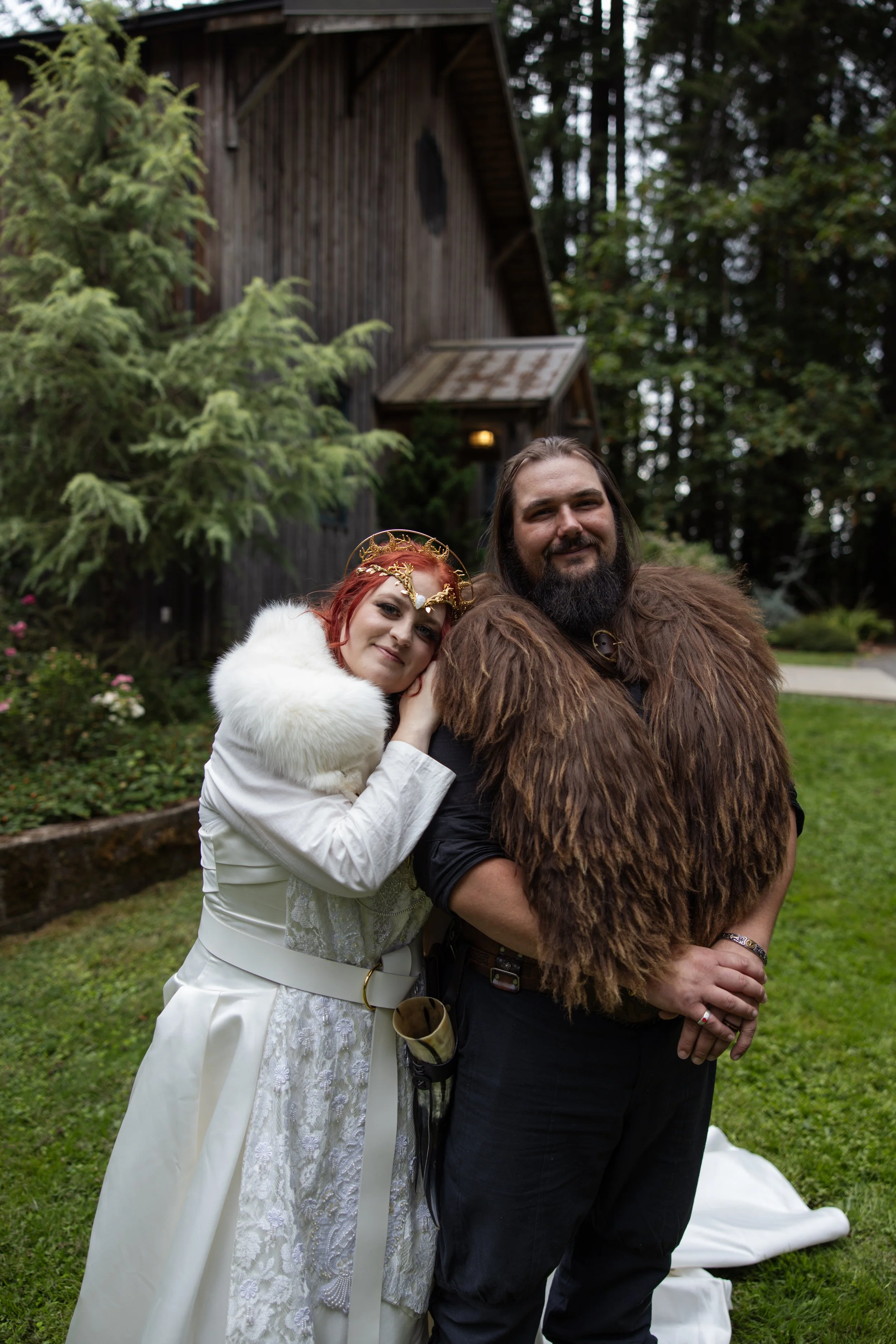 A woman with red hair in a white dress with lace details and a white fur shawl, and a man with long dark hair and beard wearing a brown furry cloak, standing outdoors in front of a wooden building with trees and shrubs in the background.