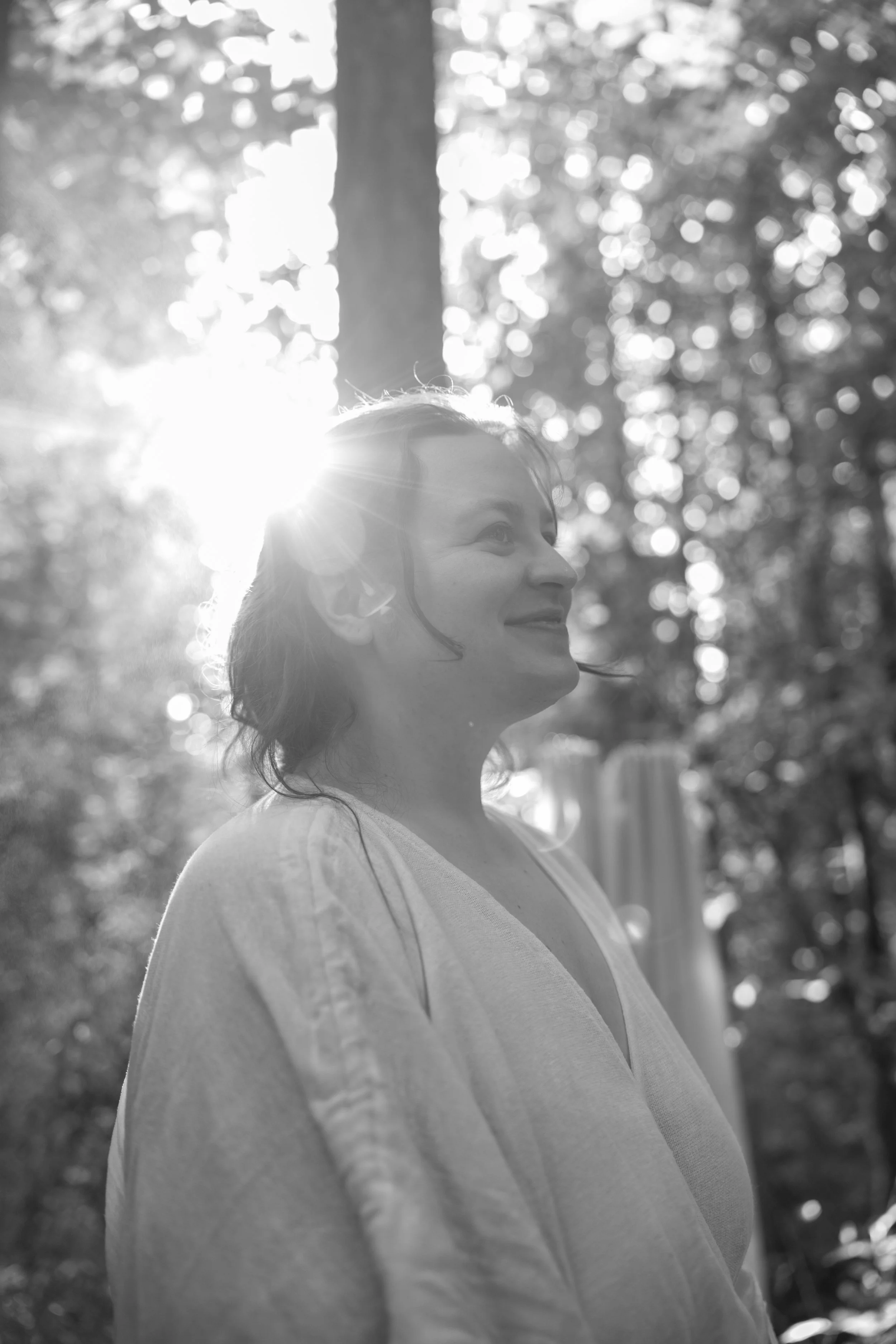 Black and white photo of a woman smiling, outdoors in a wooded area, with sunlight filtering through trees behind her.